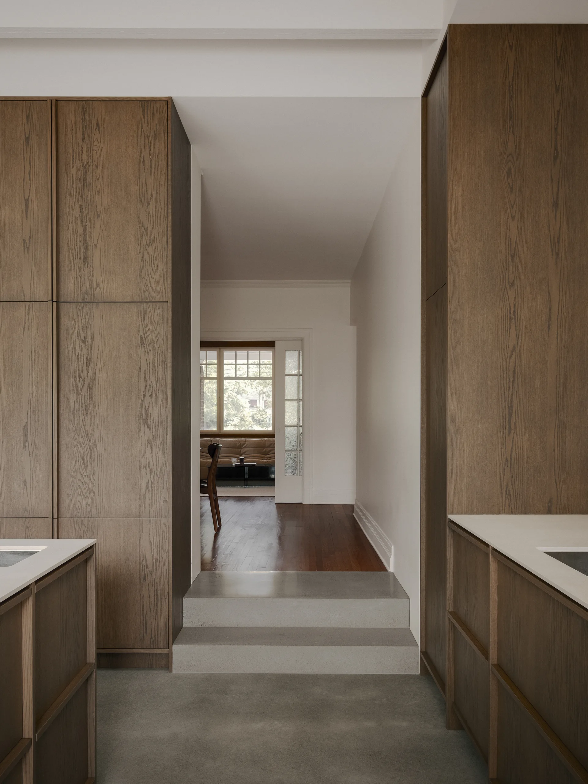 Flanked by floor-to-ceiling wood cabinetry, a grey concrete step leads from a kitchen to a hallway with wood flooring.
