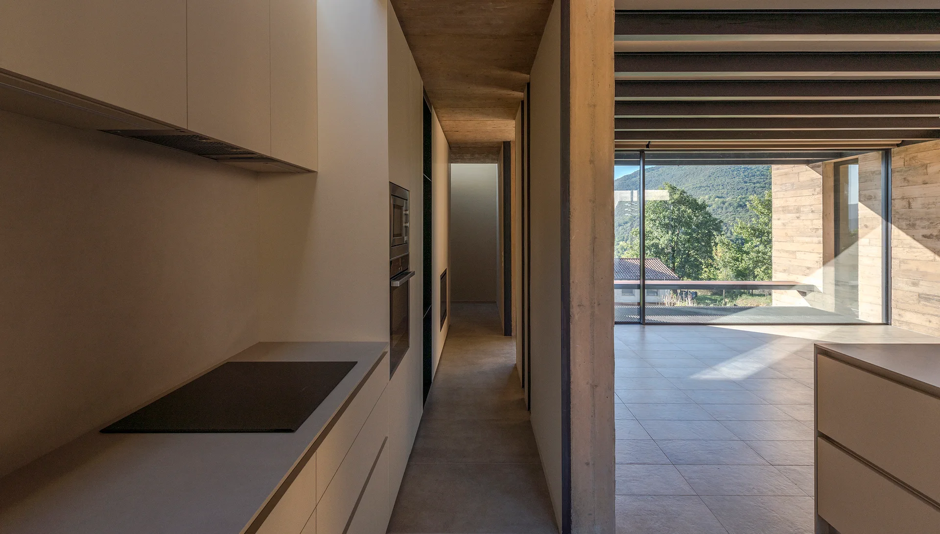 A minimalist kitchen features handleless light gray cabinets, a raw concrete pillar, and a hallway leading to a room with a large window.