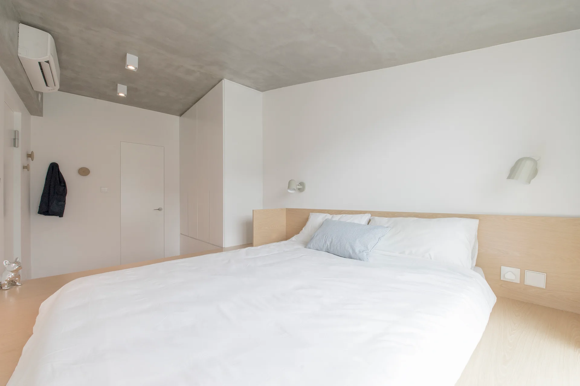 A minimalist bedroom featuring a white platform bed with a light wood headboard beneath a raw grey concrete ceiling.