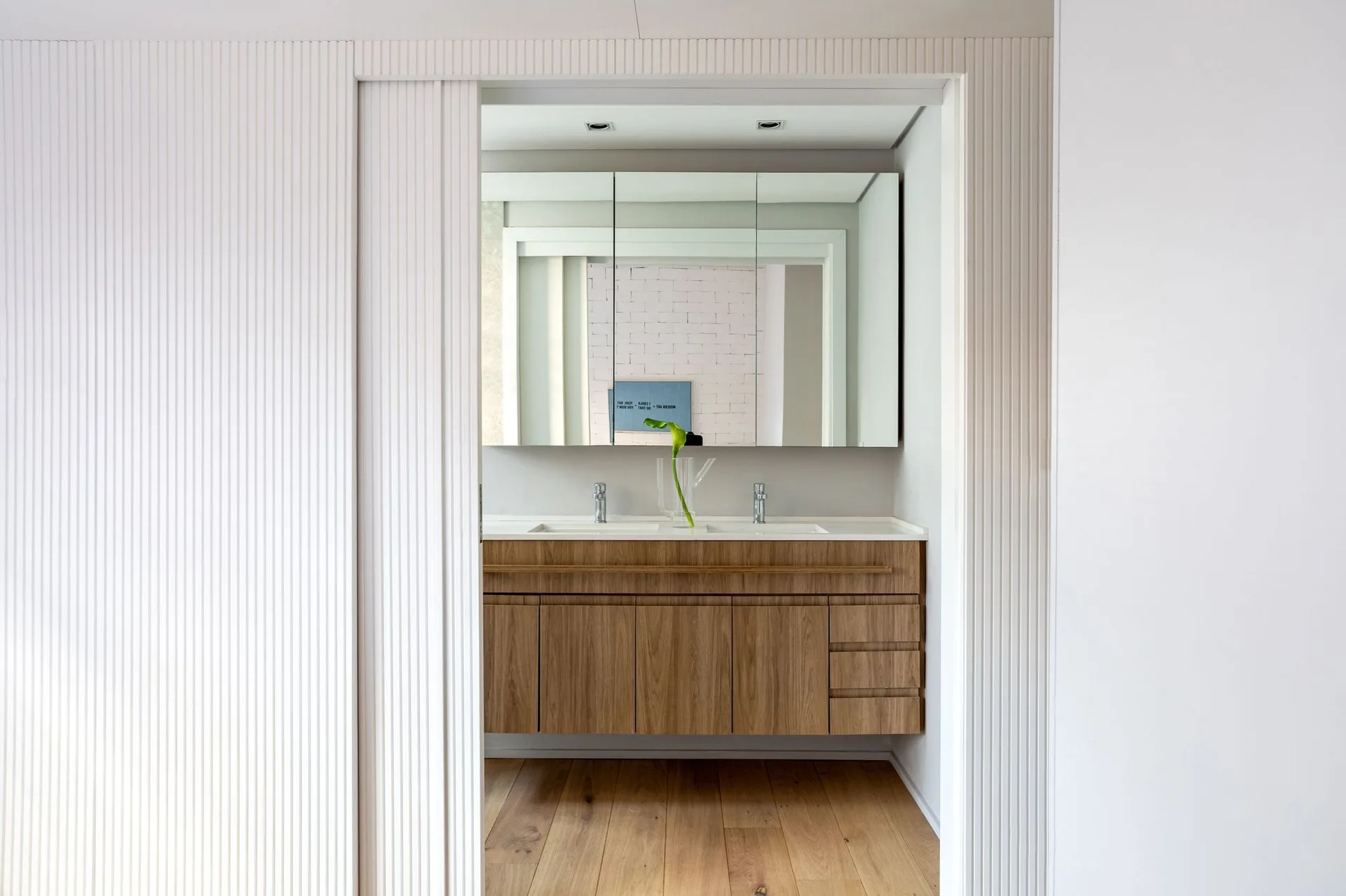 A view through a doorway of a bathroom with a floating wood vanity, white slatted wall paneling, and a mirrored cabinet.
