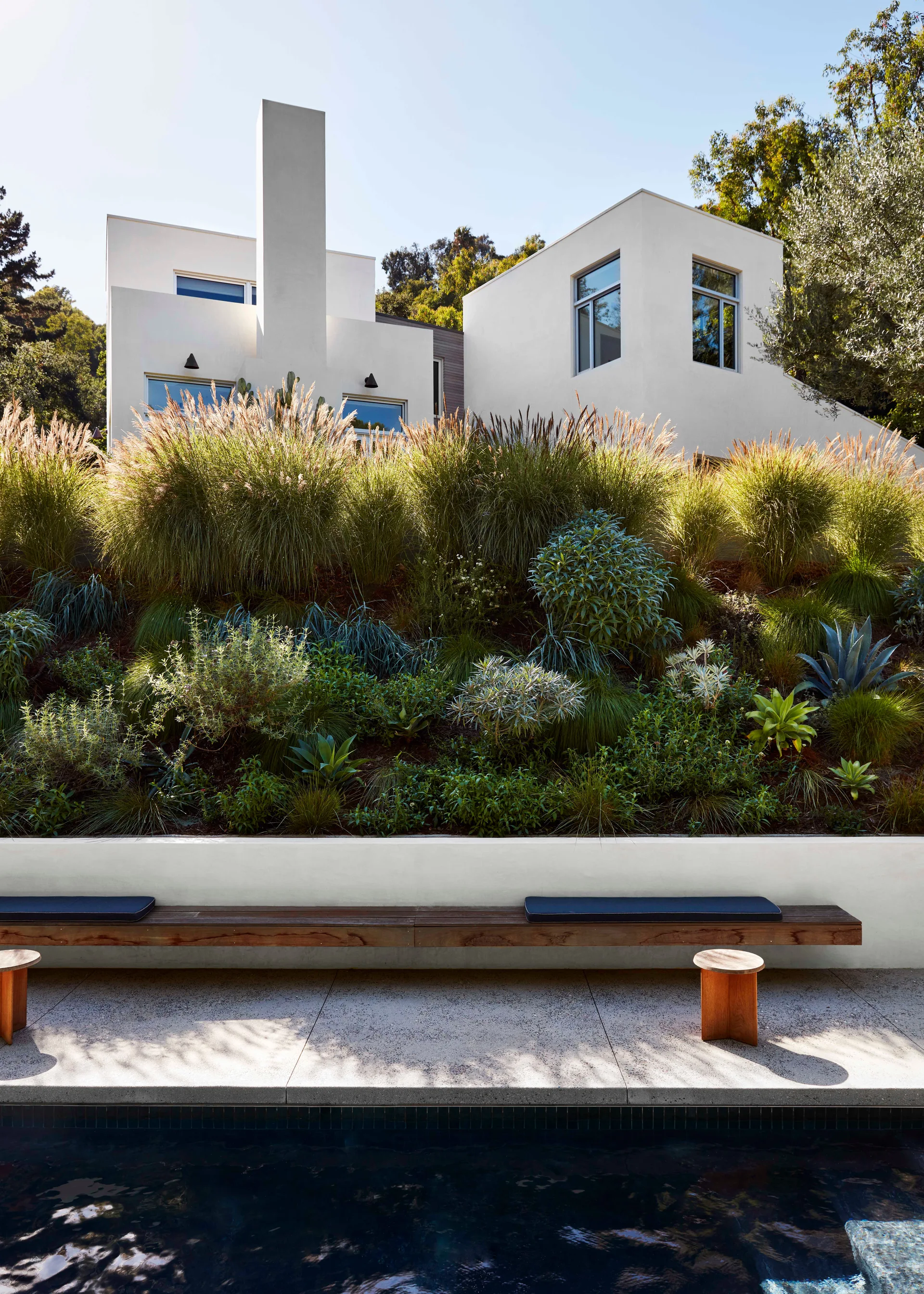 A long wooden bench with dark cushions sits beside a pool beneath a steeply terraced hillside garden and a modern white stucco house.