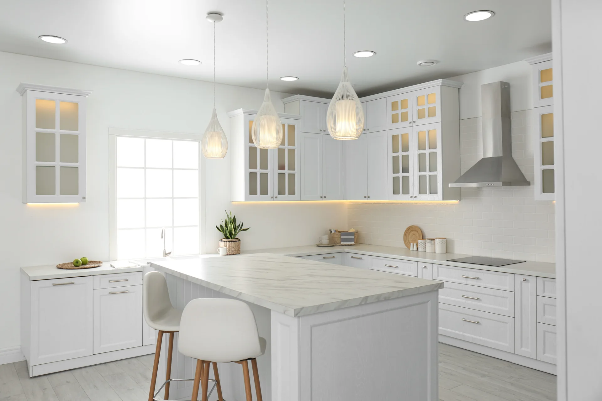 A bright white kitchen with a marble-topped island, wooden-legged stools, shaker cabinetry, and glowing teardrop pendant lights.