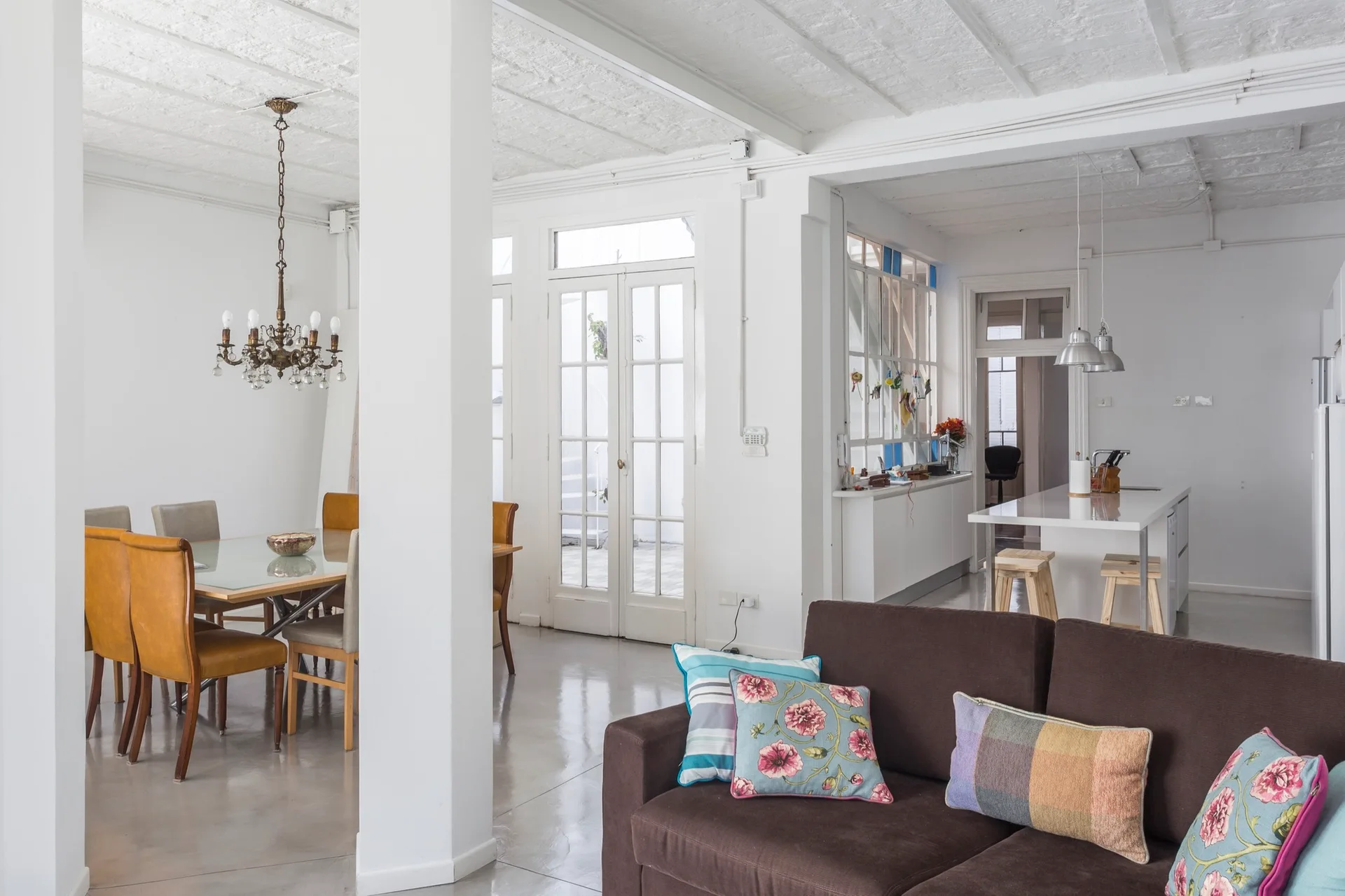 A brown sofa with colorful pillows faces an open dining area with a glass-top table, tan chairs, and white French doors in the background.