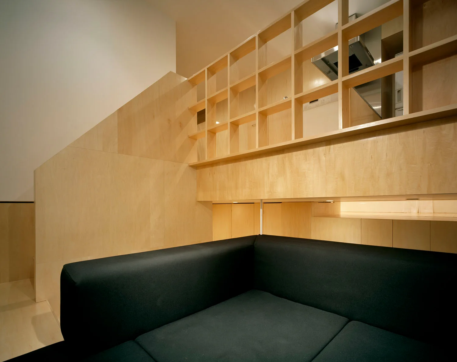 A black upholstered sofa sits below a light wood partition wall featuring an integrated grid shelf divider in a modern minimalist space.