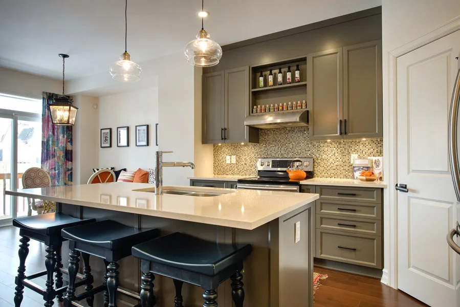 A transitional kitchen features a large island with three black bar stools, grey cabinets, a mosaic tile backsplash, and hanging glass pendant lights.