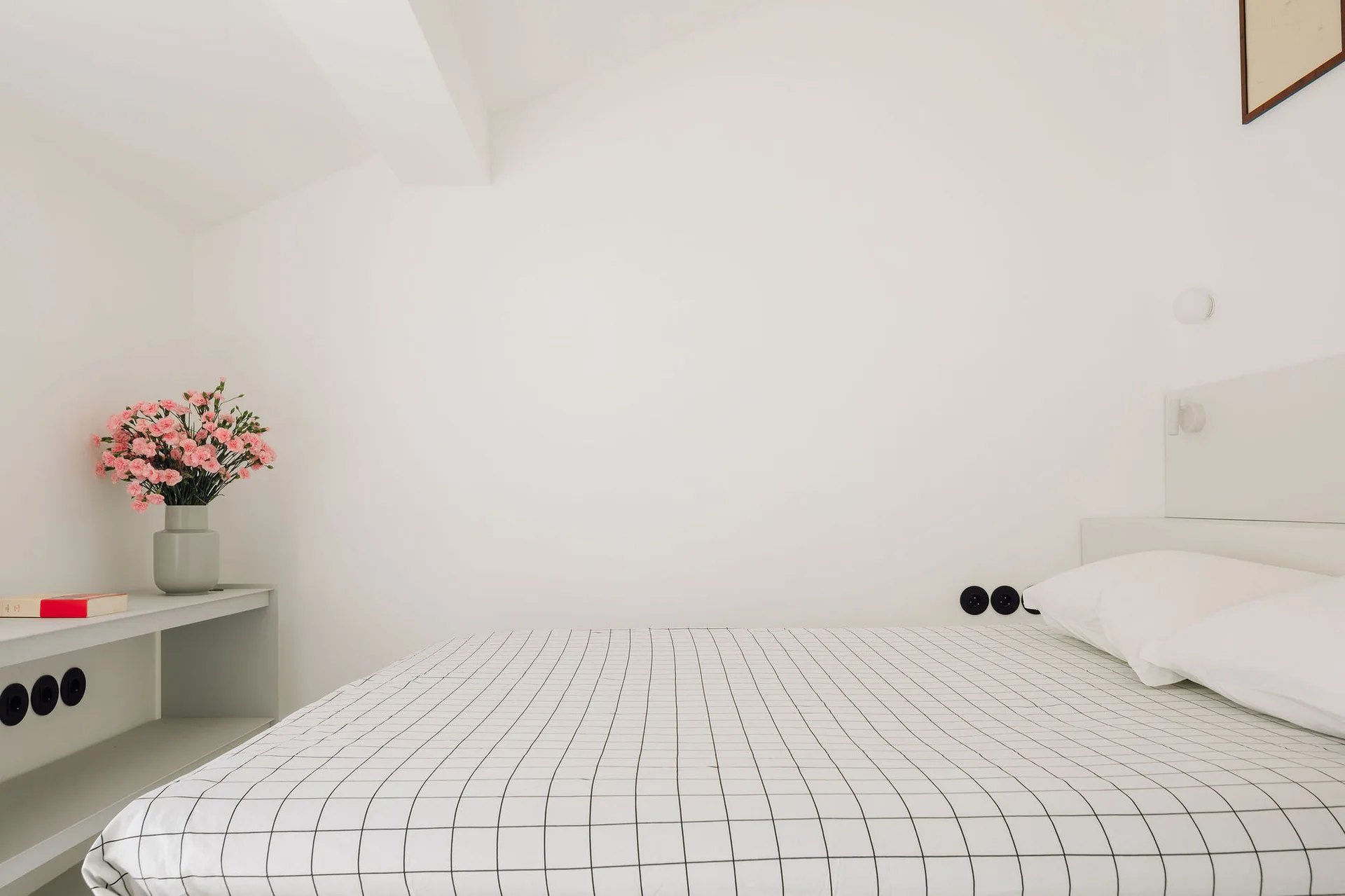 A minimalist bedroom features white bedding with a black grid pattern, a gray bedside shelf with pink flowers, and black round wall outlets.