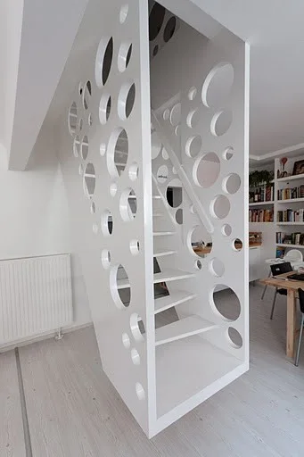 A modern white staircase featuring large and small circular cutouts on its side panels stands in a bright, open-concept living space.