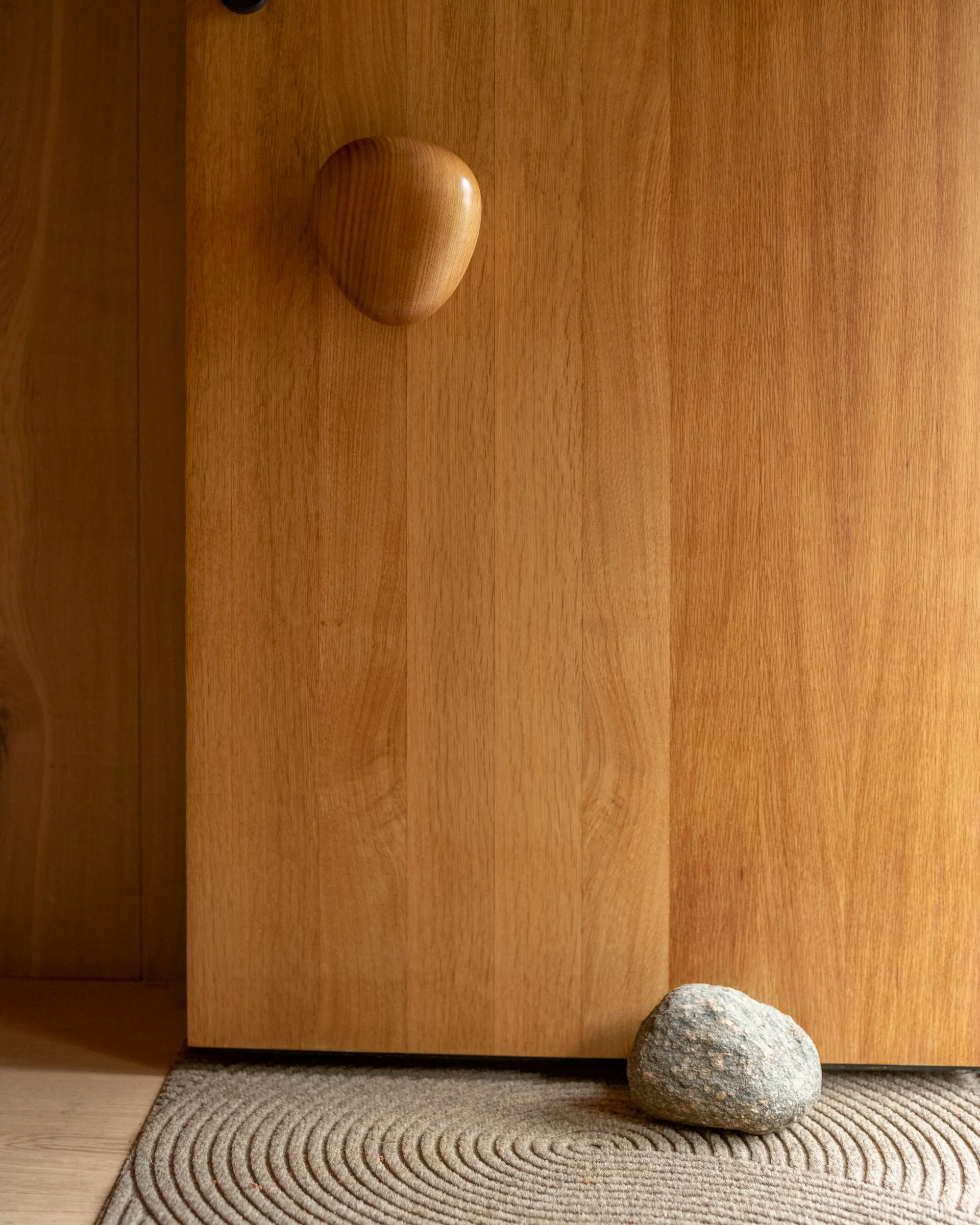 A light brown wooden door features a sculptural organic-shaped wood knob above a grey textured rug with a concentric pattern and a natural stone.