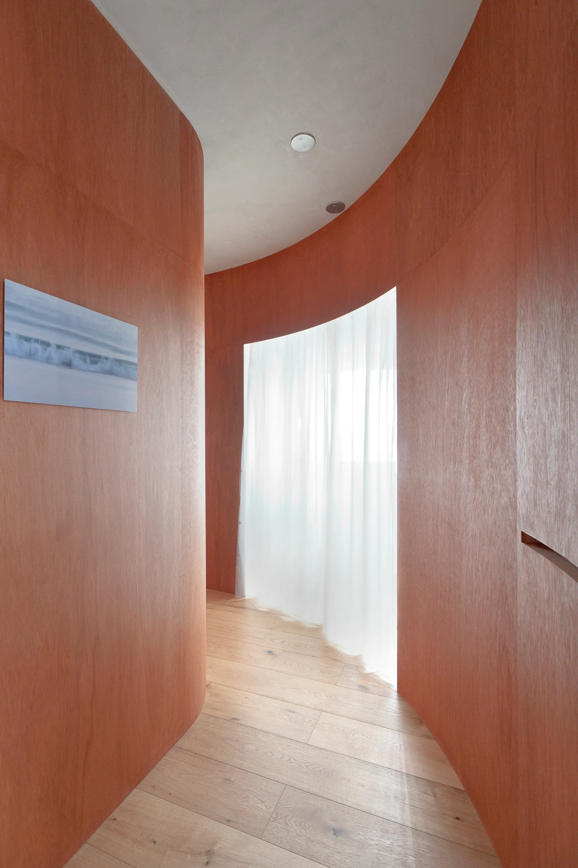 A curved hallway features warm terracotta wood-paneled walls, light wood plank floors, and a sheer white curtain filtering natural light at the end.