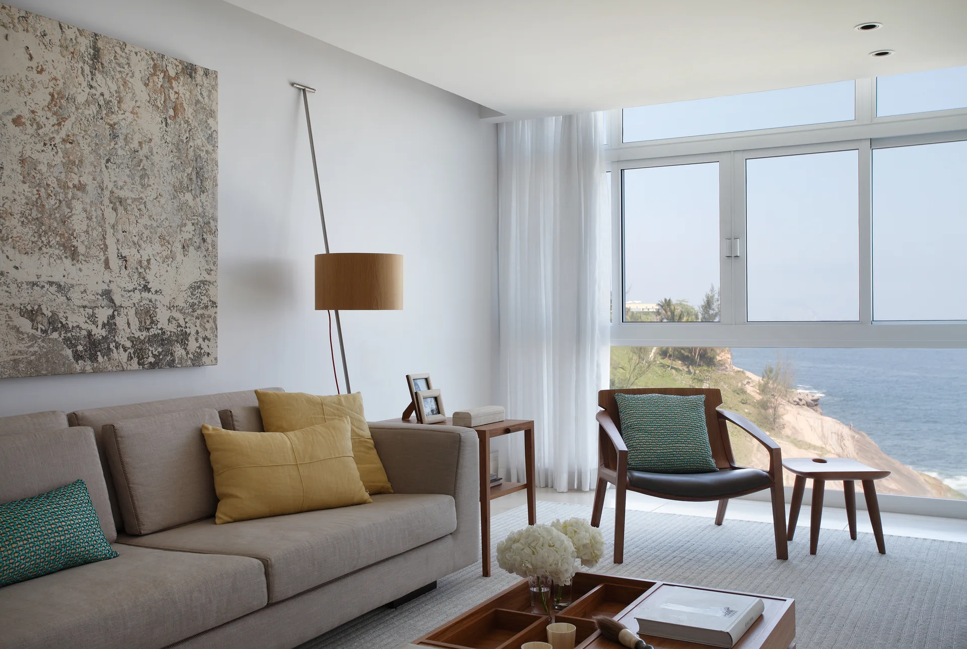 A contemporary living room with a beige sofa, wooden accent chair, and a large window looking out at a rocky ocean coastline.