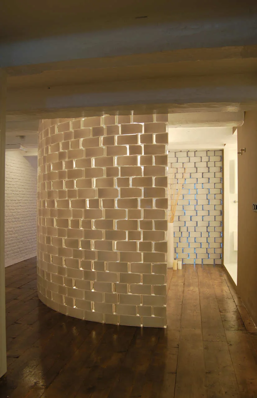 A curved partition wall made of translucent glass blocks stands on dark wood floorboards beneath white exposed ceiling beams.