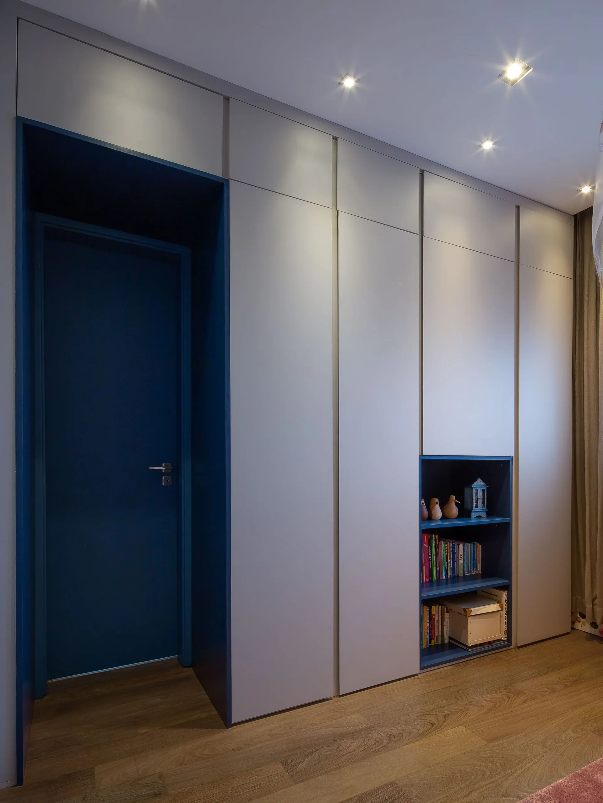 A floor-to-ceiling light gray custom storage unit featuring a built-in deep blue door frame and an inset open bookshelf on wood flooring.
