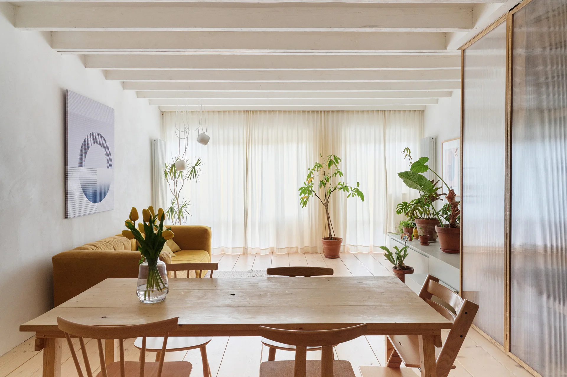 An open-concept dining area with a wooden table, chairs, white ceiling beams, and a mustard yellow sofa near sheer window curtains.