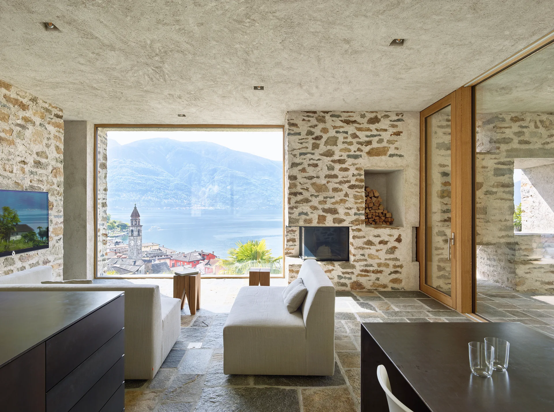 A rustic contemporary living room features exposed stone walls, light grey sofas, and a large panoramic window showing a lake and mountain landscape.
