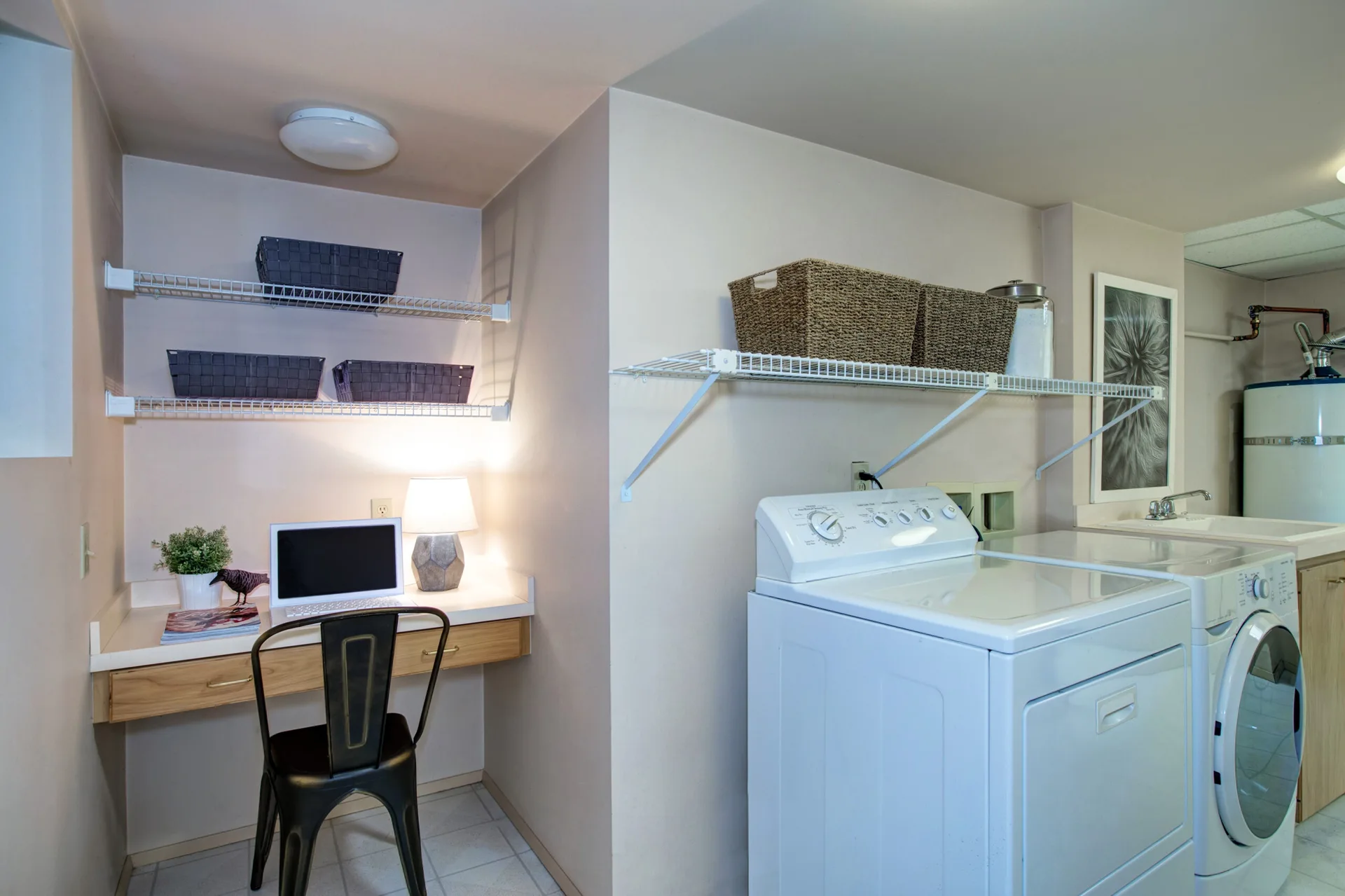A functional laundry room featuring white washing appliances and wire shelving next to a built-in wooden desk with a black metal chair.