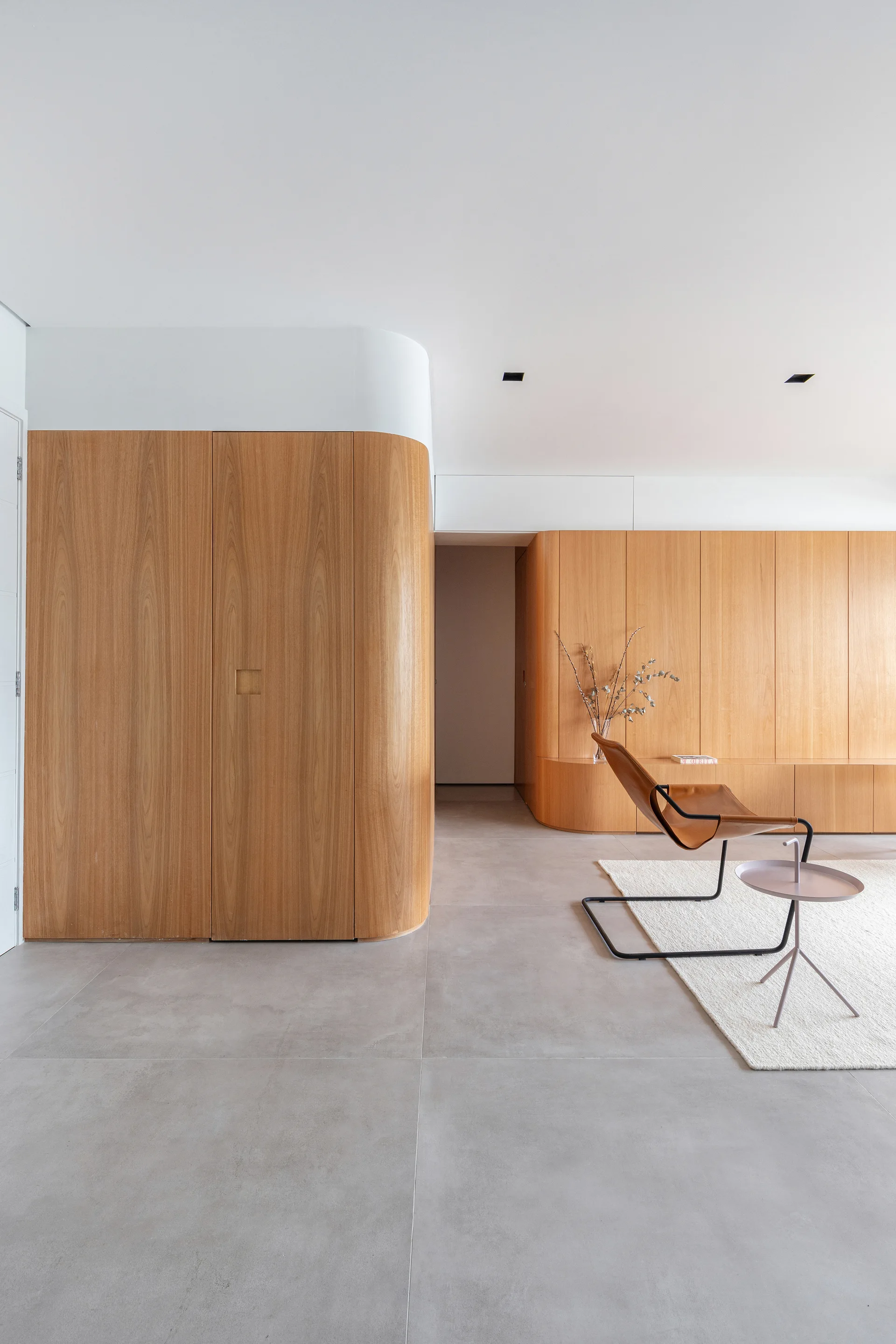 Minimalist living room featuring curved light wood paneling, a tan leather sling chair on a cream rug, and grey tile floors.