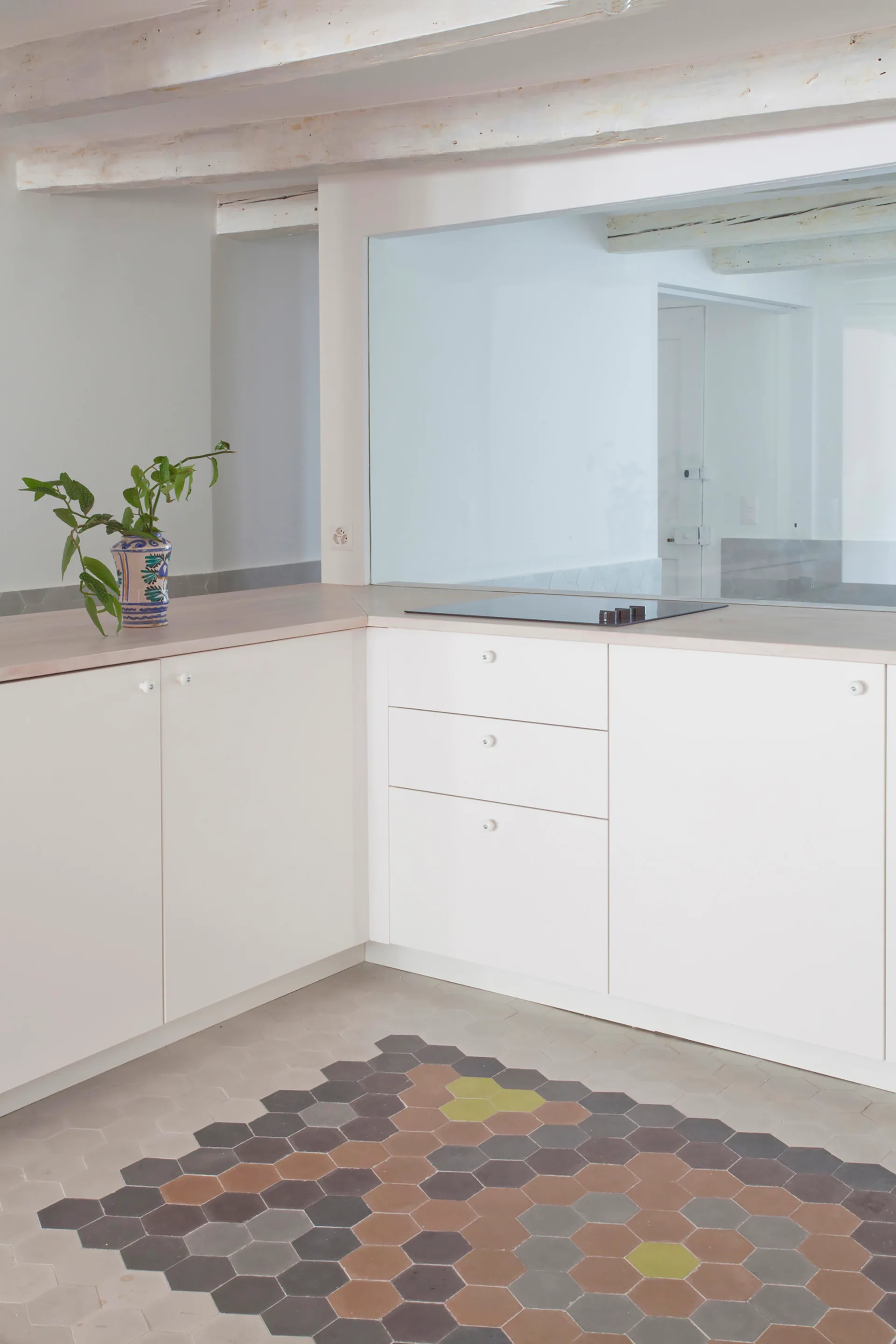 A minimalist white kitchen features flat-front cabinets, a light wood countertop with a cooktop, and a patterned hexagonal tile floor.