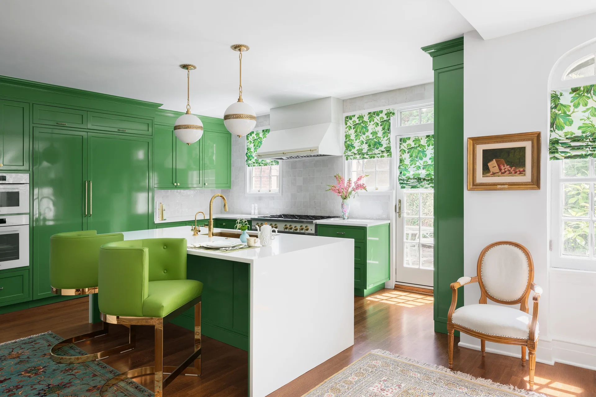 A bright kitchen features glossy green cabinetry, a white central island with green barstools, botanical shades, and brass pendant lights.
