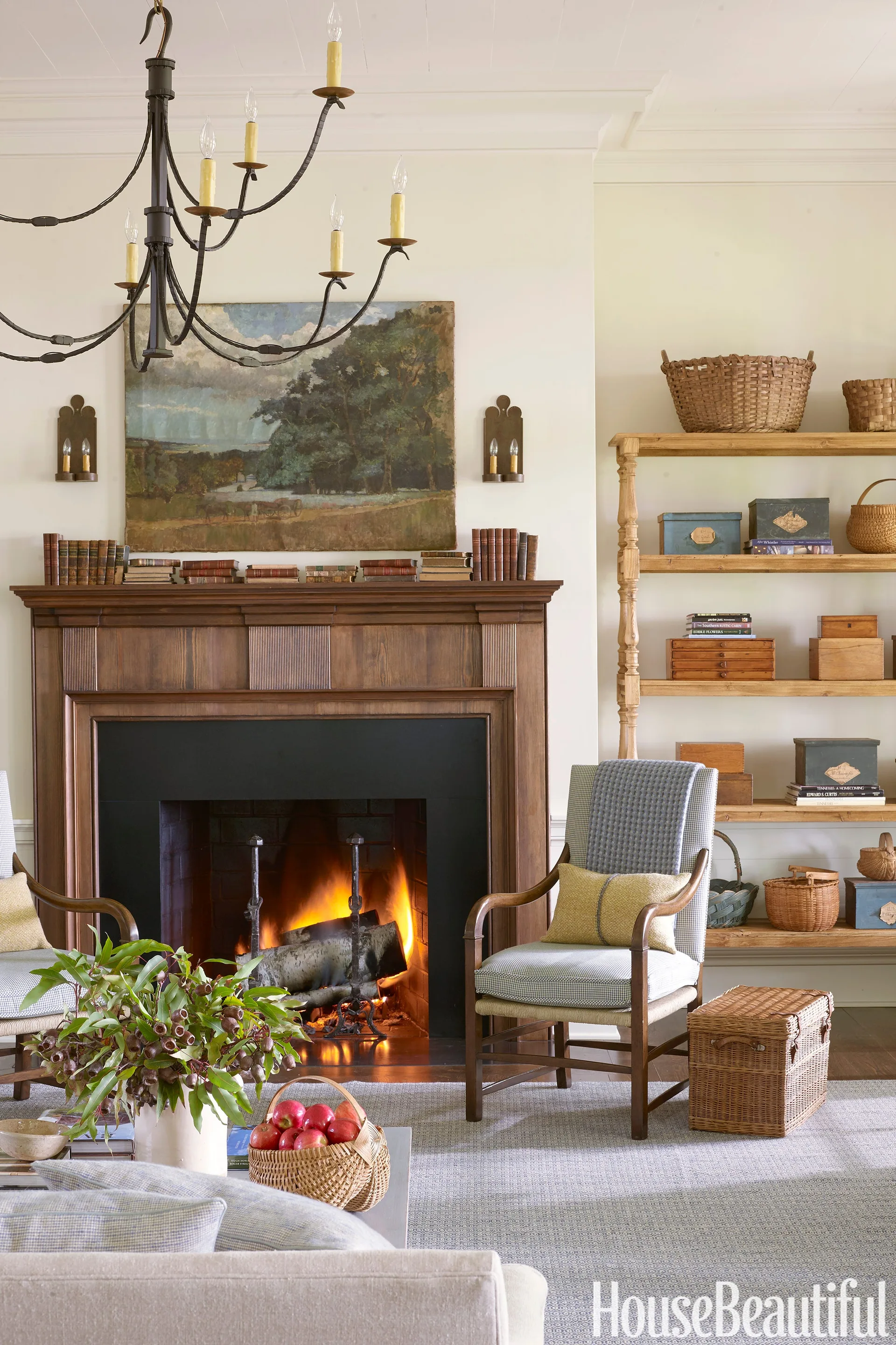 A cozy living room features a burning fireplace with a dark wood mantel, flanked by a patterned armchair and a tall wooden bookshelf filled with baskets.