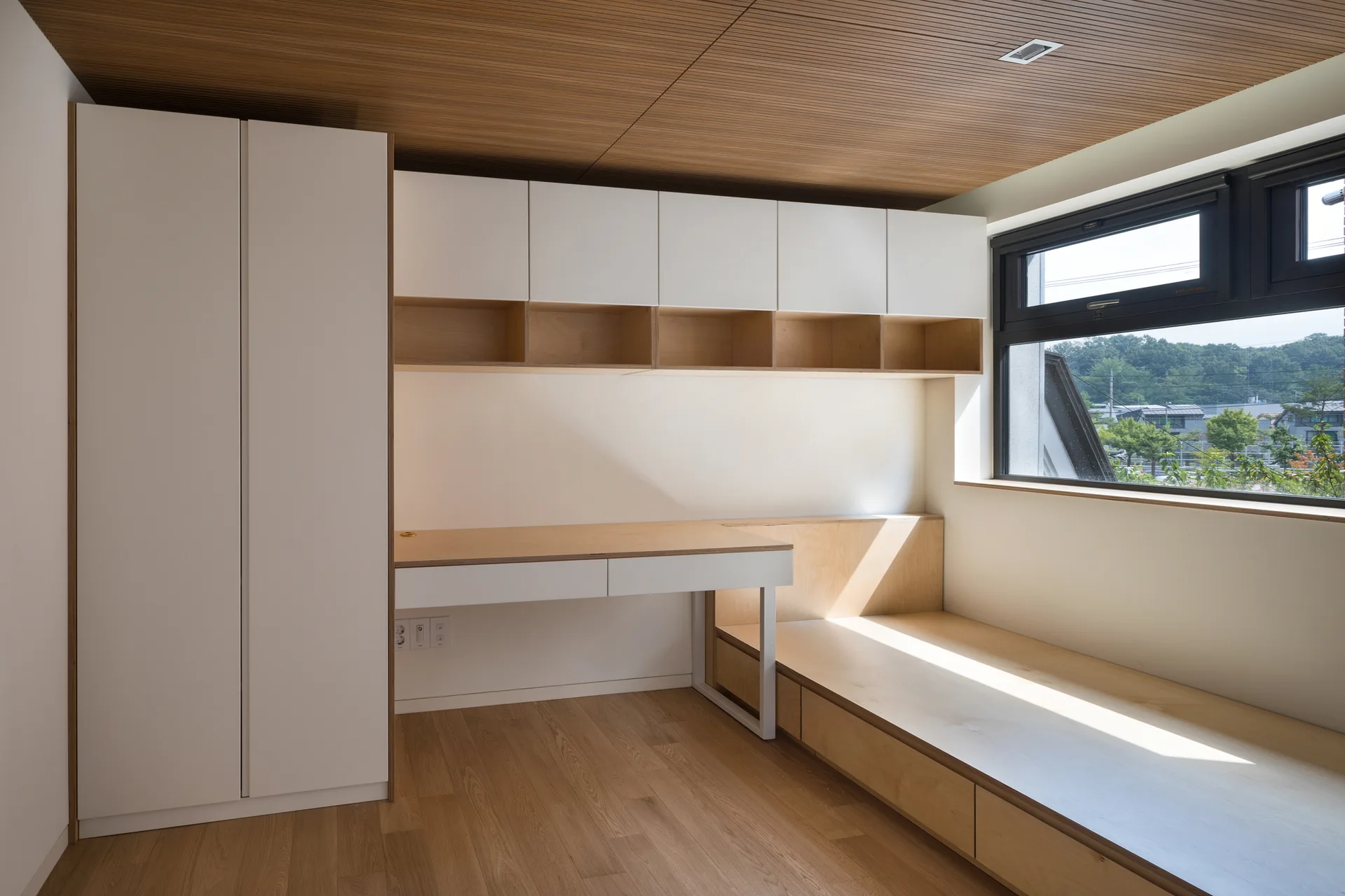 A minimalist room featuring a built-in light wood platform bed, matching desk, white cabinets, and a slatted wood ceiling.