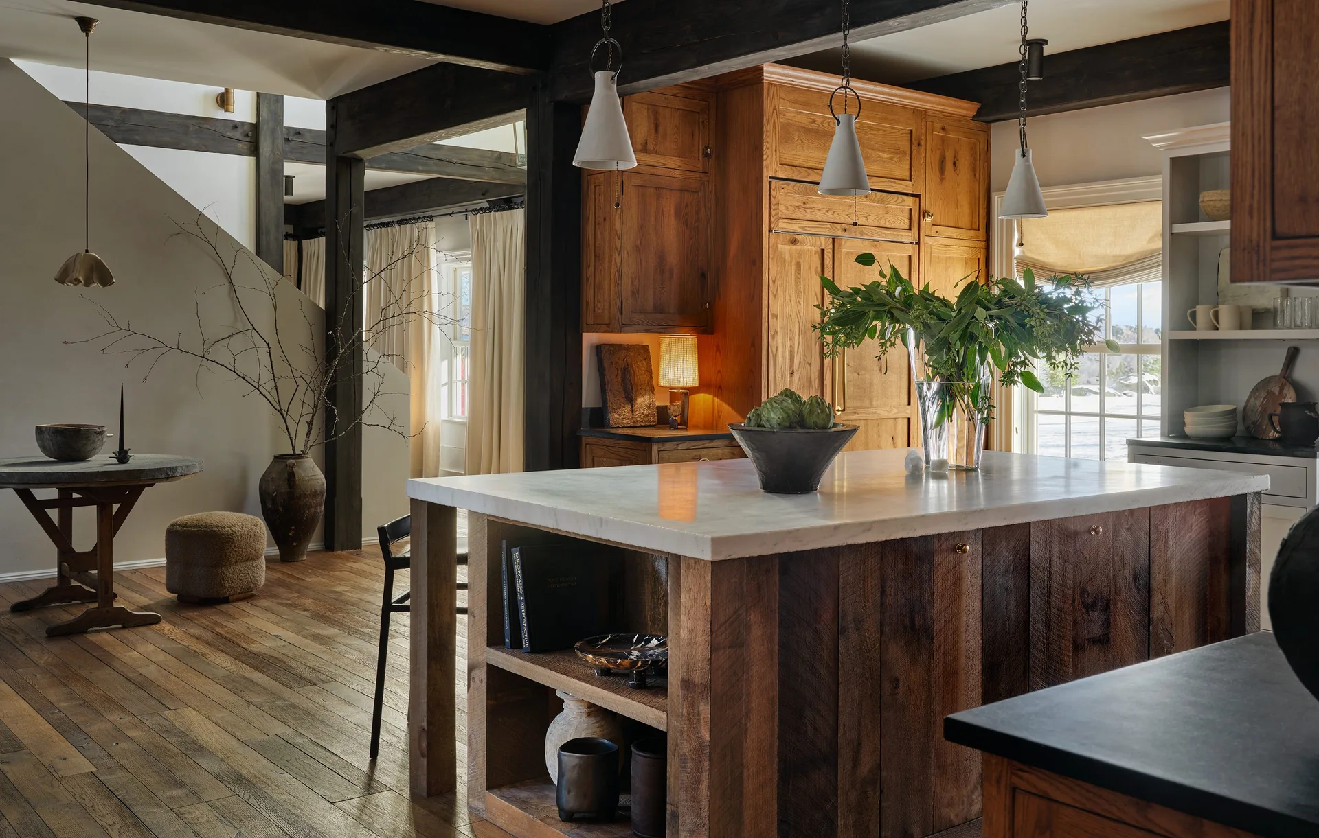 A rustic kitchen showcasing a wood island with a white marble top, dark exposed ceiling beams, and warm wood cabinetry.