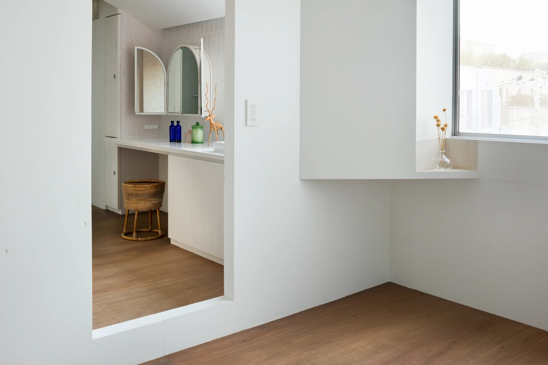 A rectangular wall cutout frames a white minimalist bathroom vanity with an arched mirror, a rattan stool, and natural wood floors.