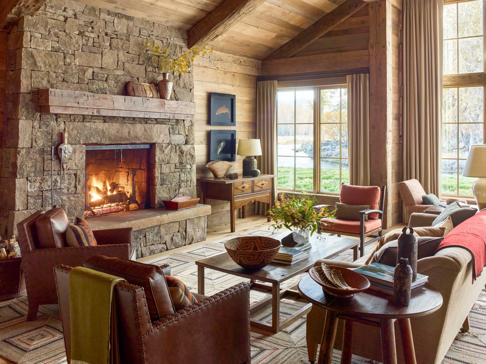 A rustic living room featuring a large stone fireplace, horizontal wood plank walls, leather armchairs, and a vaulted ceiling with exposed beams.