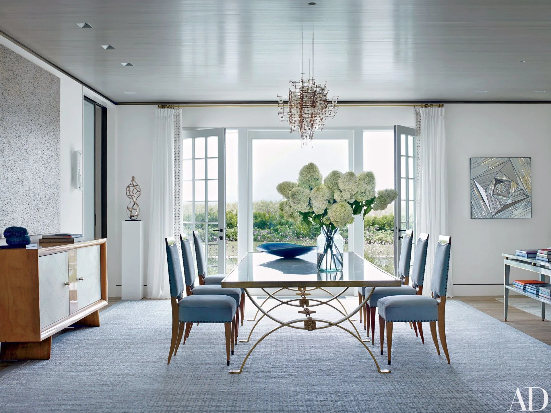 A bright dining room features a glass-top dining table with blue upholstered chairs, a modern chandelier, and open French doors leading to a garden.