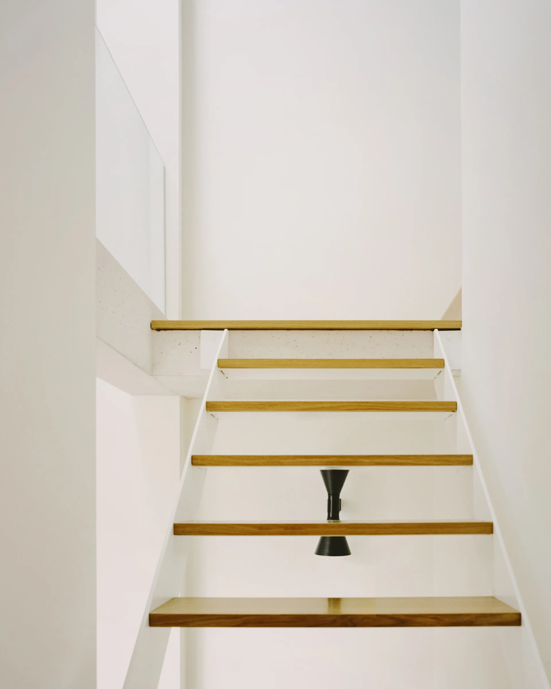 A minimalist staircase features open light wood treads, white metal stringers, and a small black sconce mounted on the wall behind the steps.