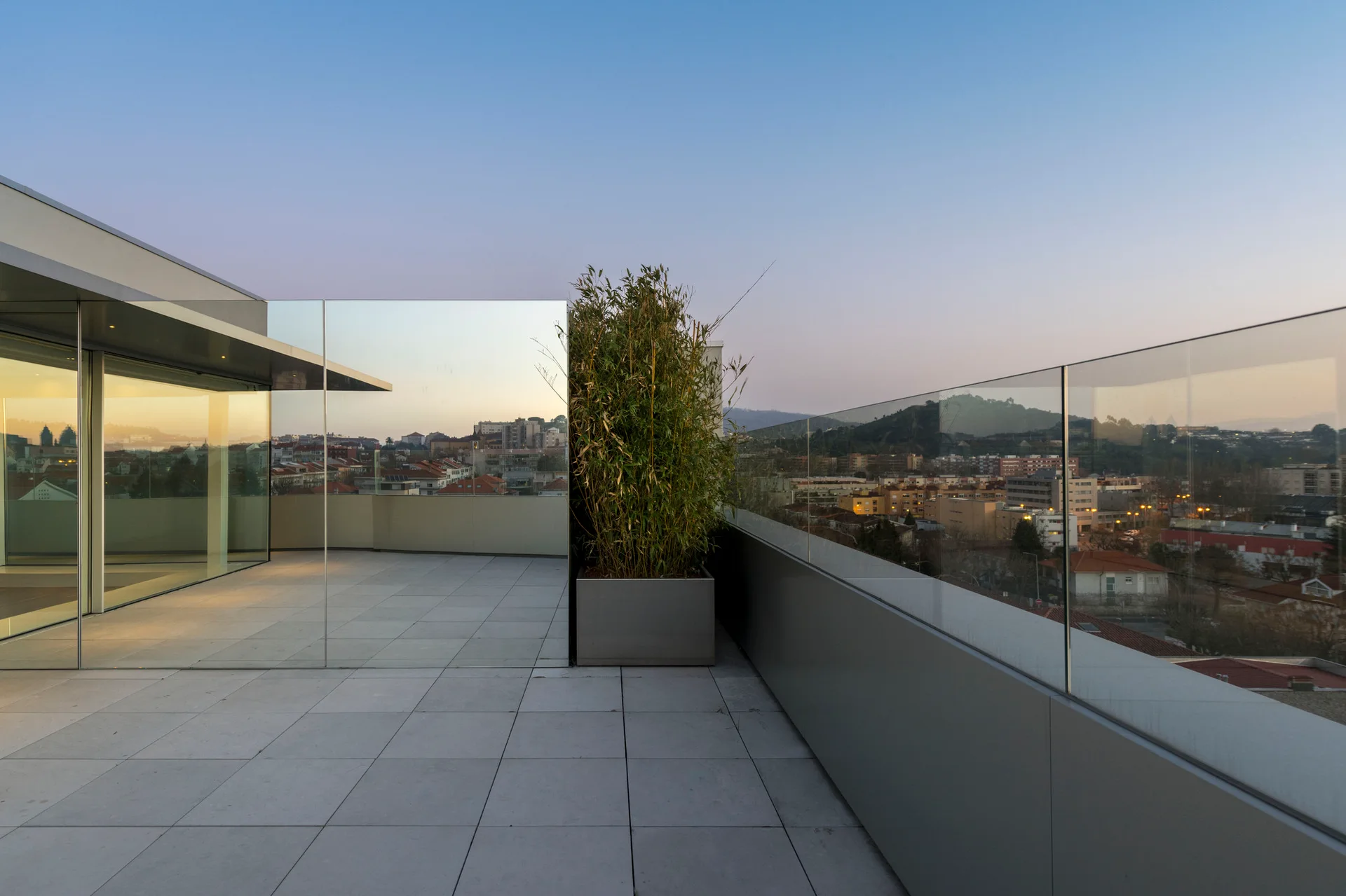 A modern outdoor terrace with grey stone tile flooring, a frameless glass railing, and a silver planter containing a tall bamboo plant.