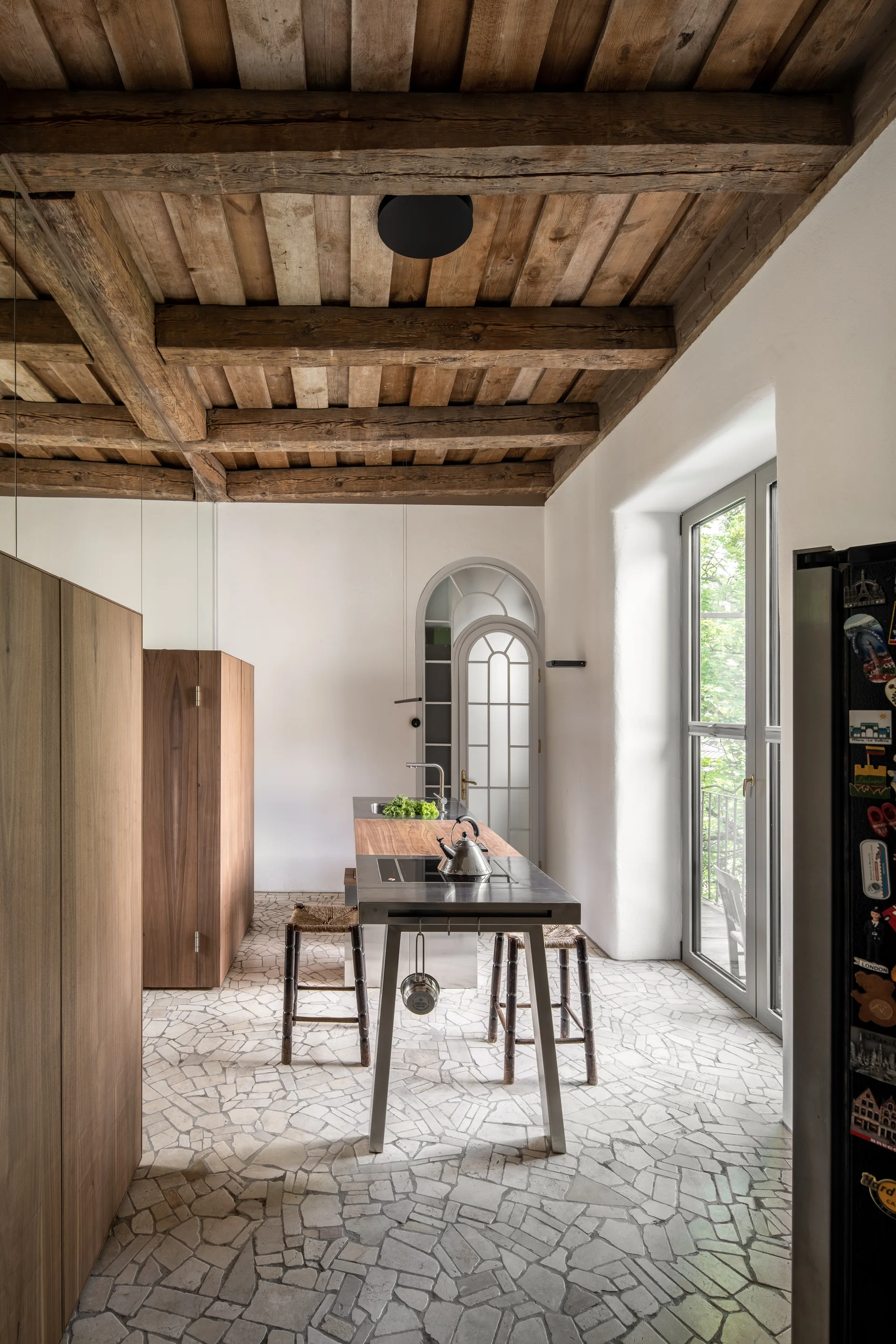 A rustic kitchen features exposed wooden ceiling beams, an irregular stone tiled floor, and a modern metal and wood island with stools.