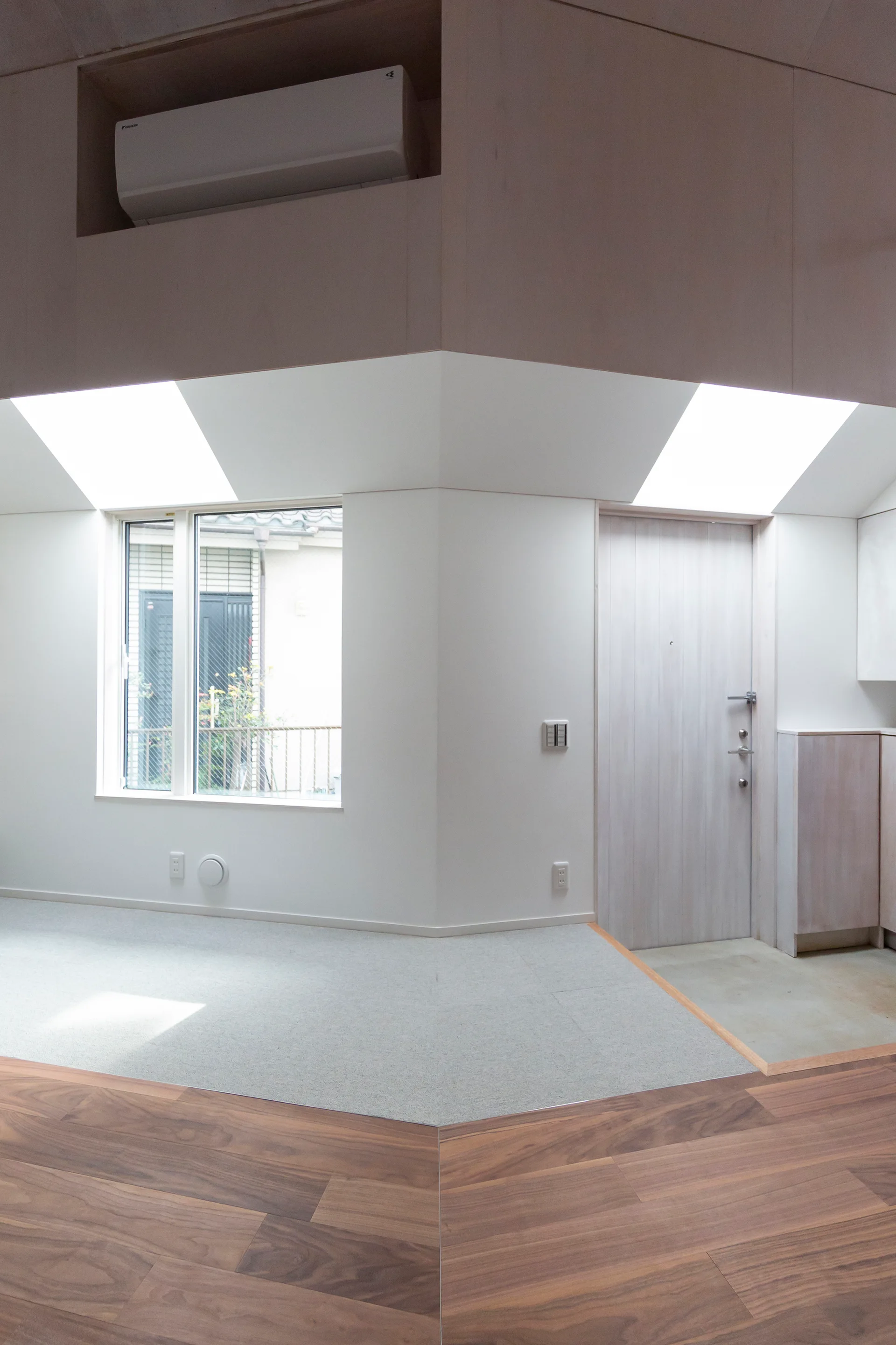 A minimalist residential entryway featuring a light wood door, angular skylights on a sloped ceiling, and mixed wood and concrete flooring.