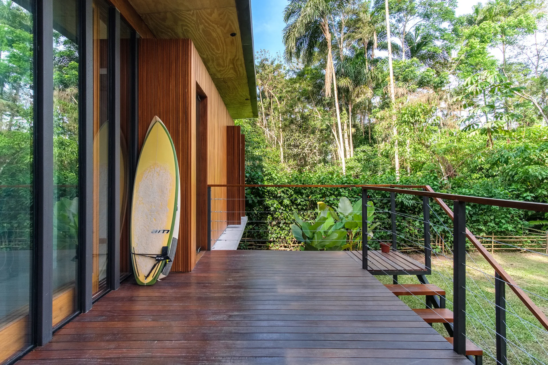 A wooden deck with vertical slat siding features a yellow surfboard leaning against the wall, a cable railing, and a background of tropical green trees.