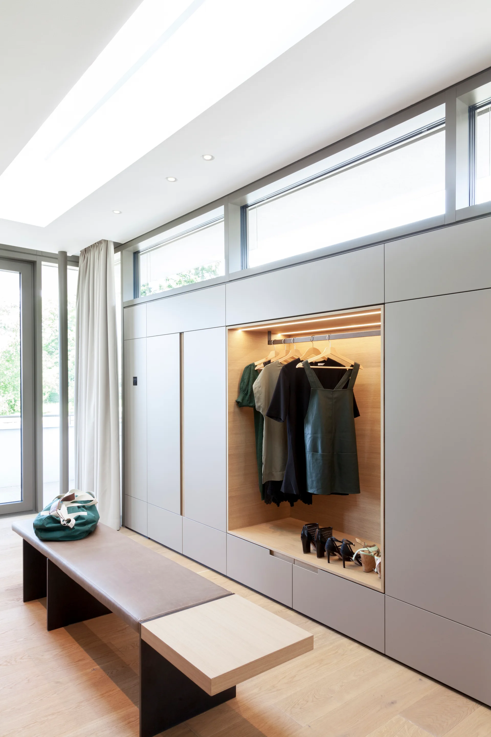 A modern dressing room with matte grey built-in closets, an open wooden wardrobe niche, and a leather-topped bench on light oak floors.