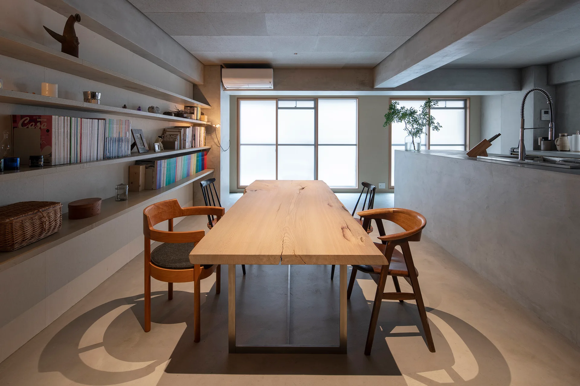 A long live-edge wooden dining table with chairs sits in an industrial room featuring concrete walls, built-in concrete shelving, and frosted windows.