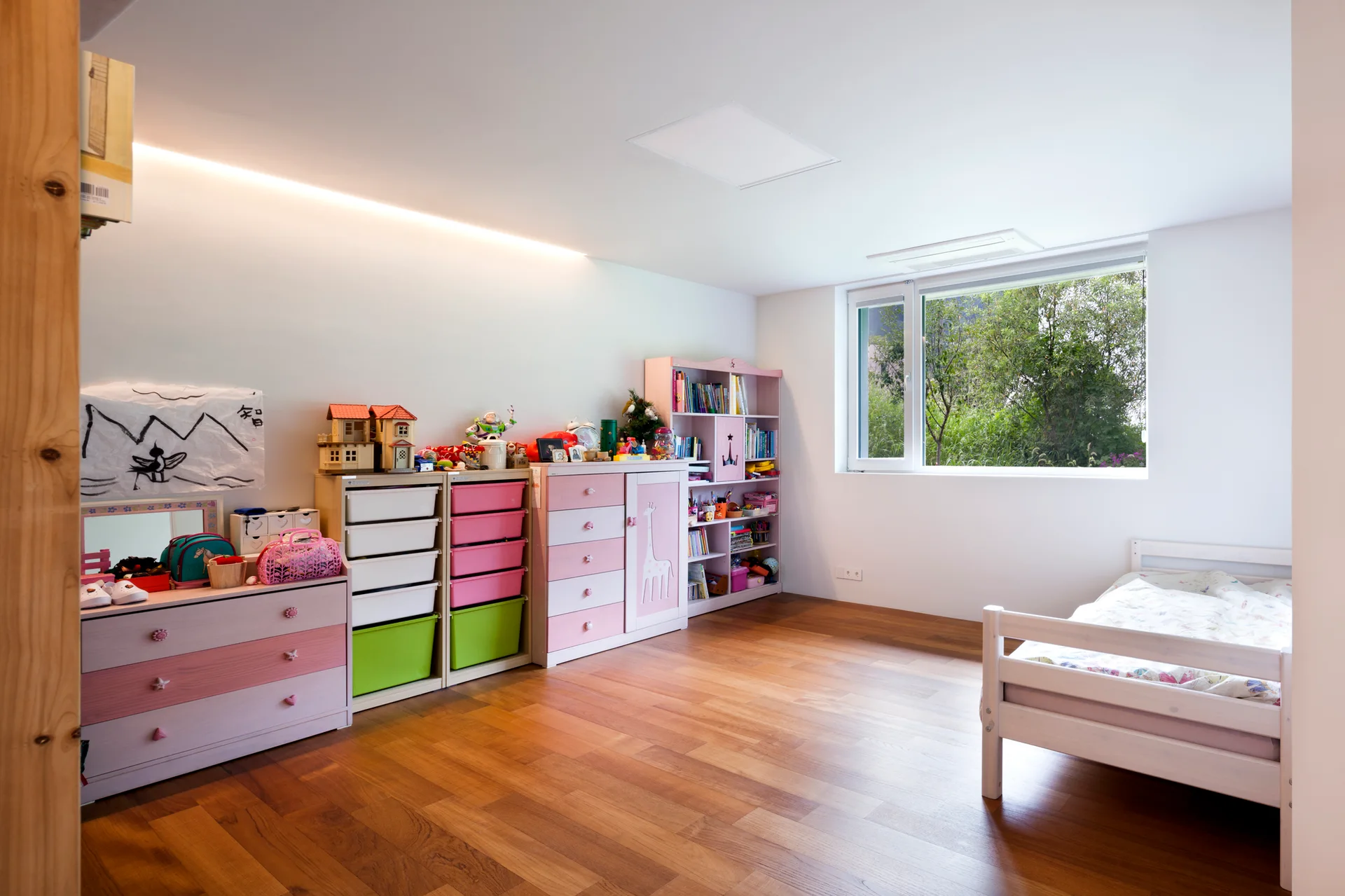 A bright children's bedroom featuring pink and white toy storage units, a simple white wooden bed, and warm wood flooring.