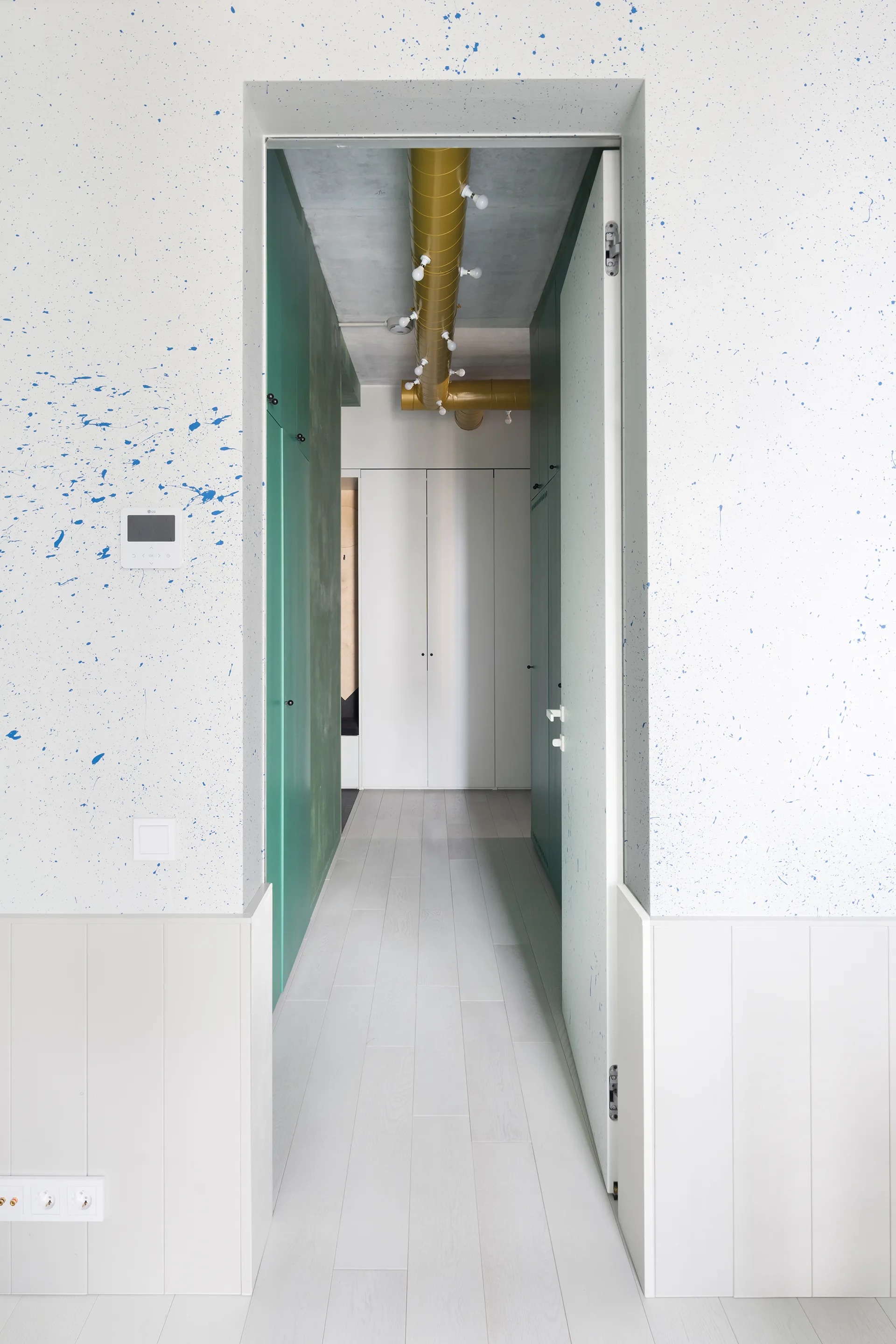 A modern hallway features white walls with blue paint splatter, lower white wainscoting, green cabinetry, and a bright yellow ceiling fixture.