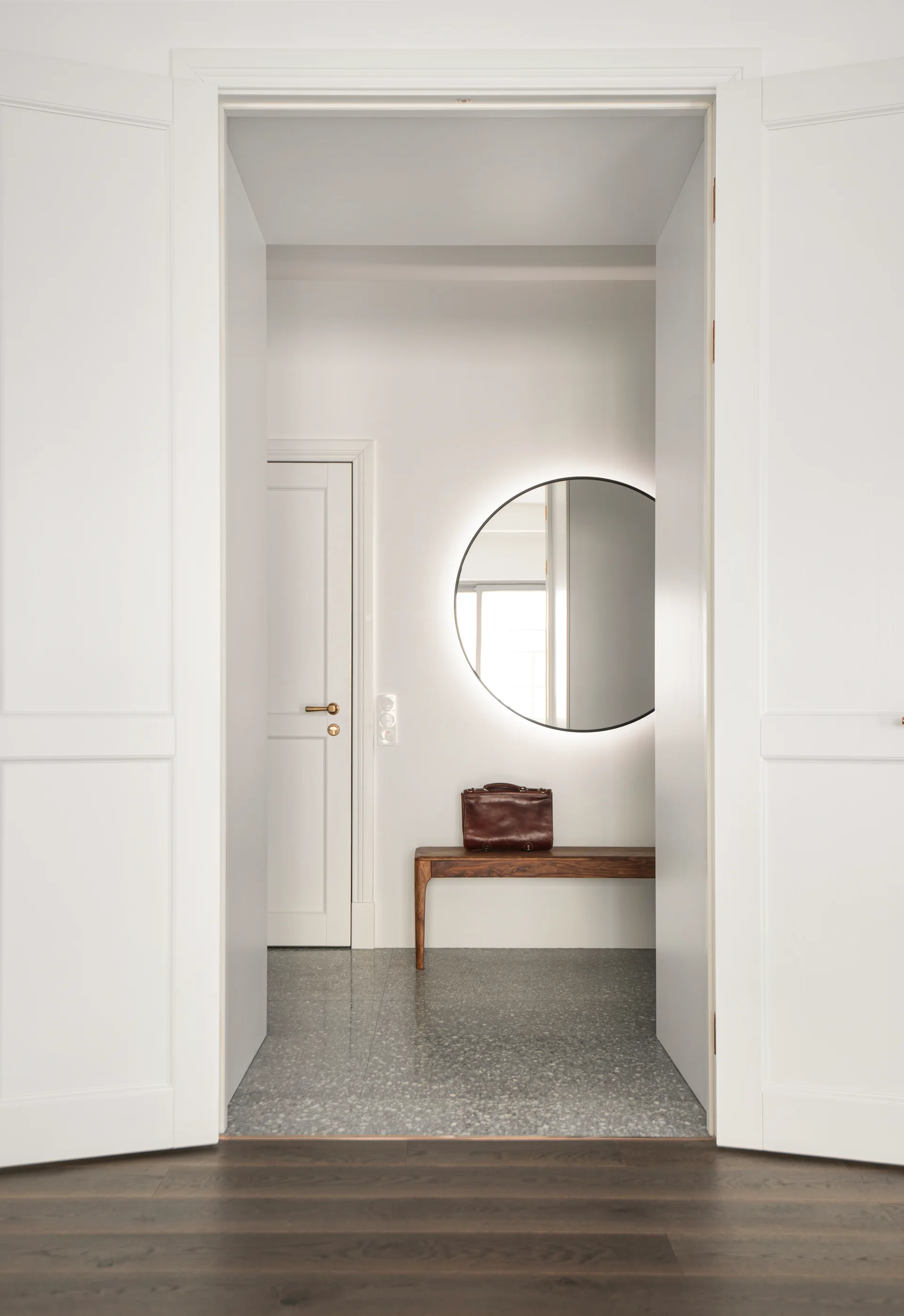 A minimalist entryway with grey terrazzo flooring, a wooden bench, and a backlit round mirror is viewed through open white double doors.