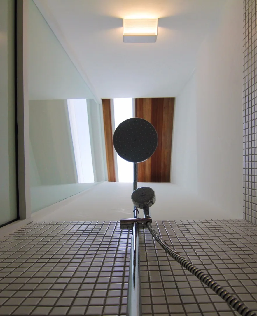 A low-angle view of a chrome shower head against white mosaic tile walls and a high ceiling with a wood-plank skylight.