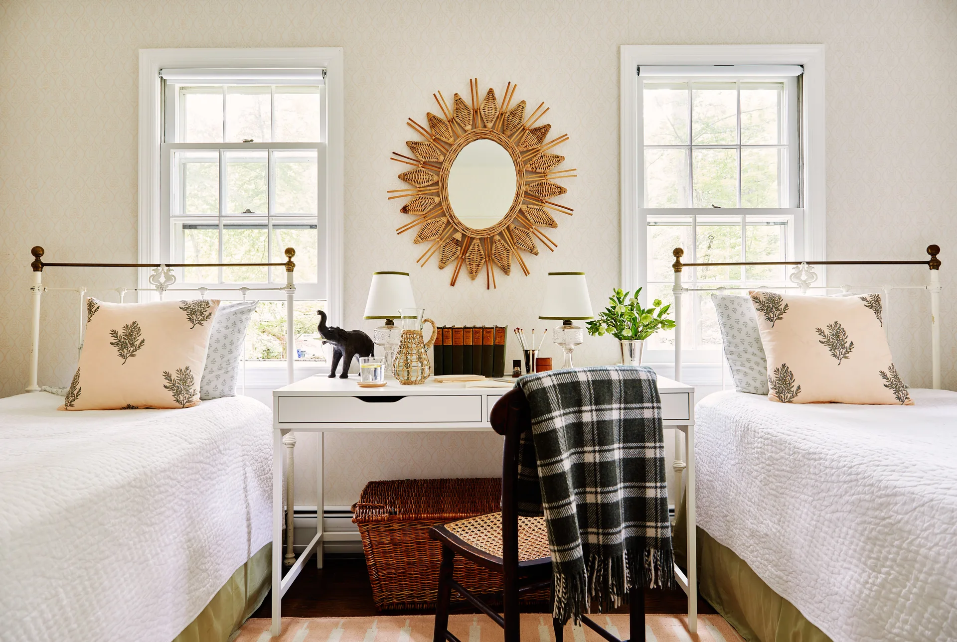 Two white twin beds sit symmetrically on either side of a white writing desk with a wicker sunburst mirror hanging on the patterned wallpaper above.