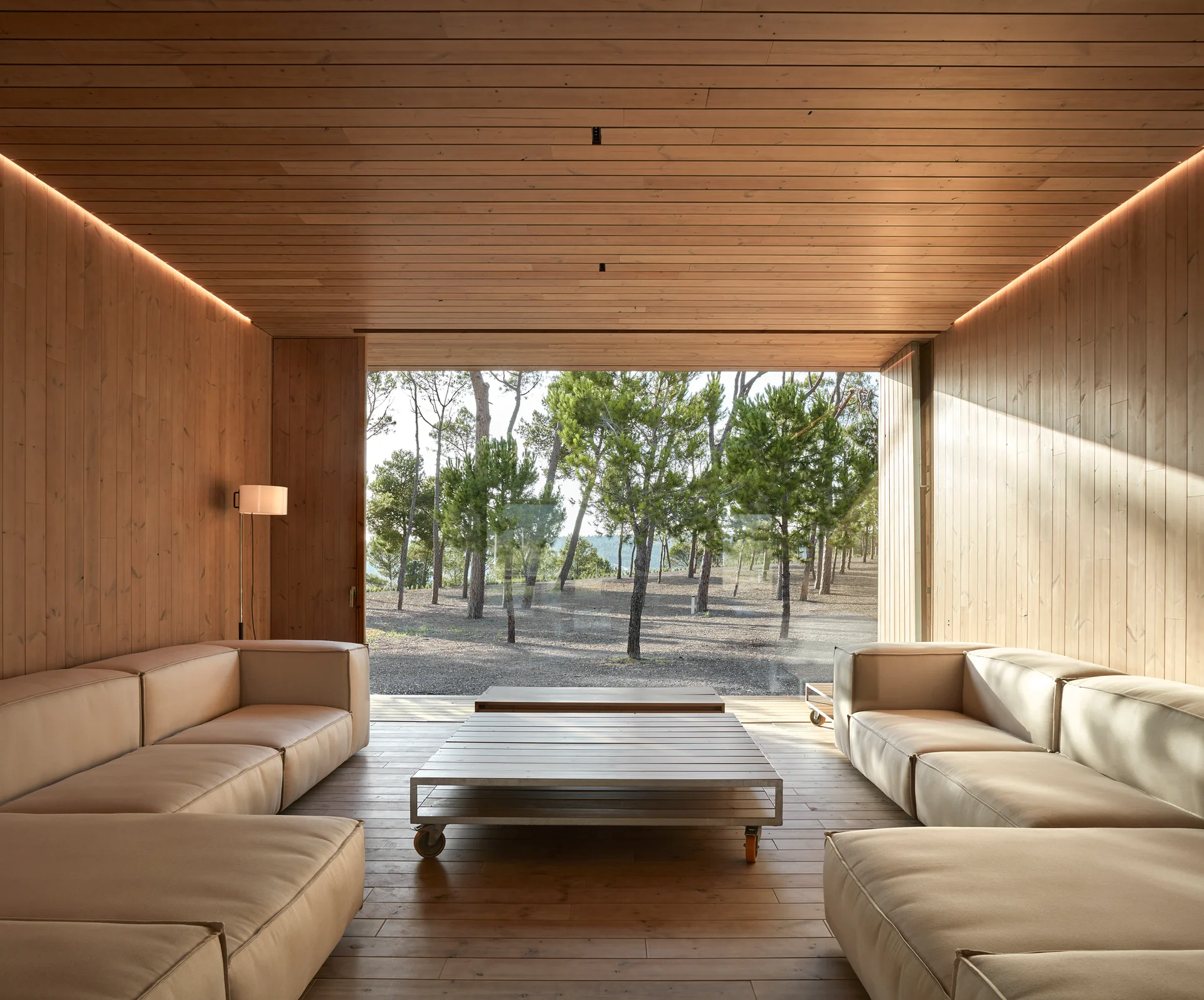 A symmetrical living room featuring wood-paneled surfaces, two beige modular sofas, and a large window looking out onto a pine forest.