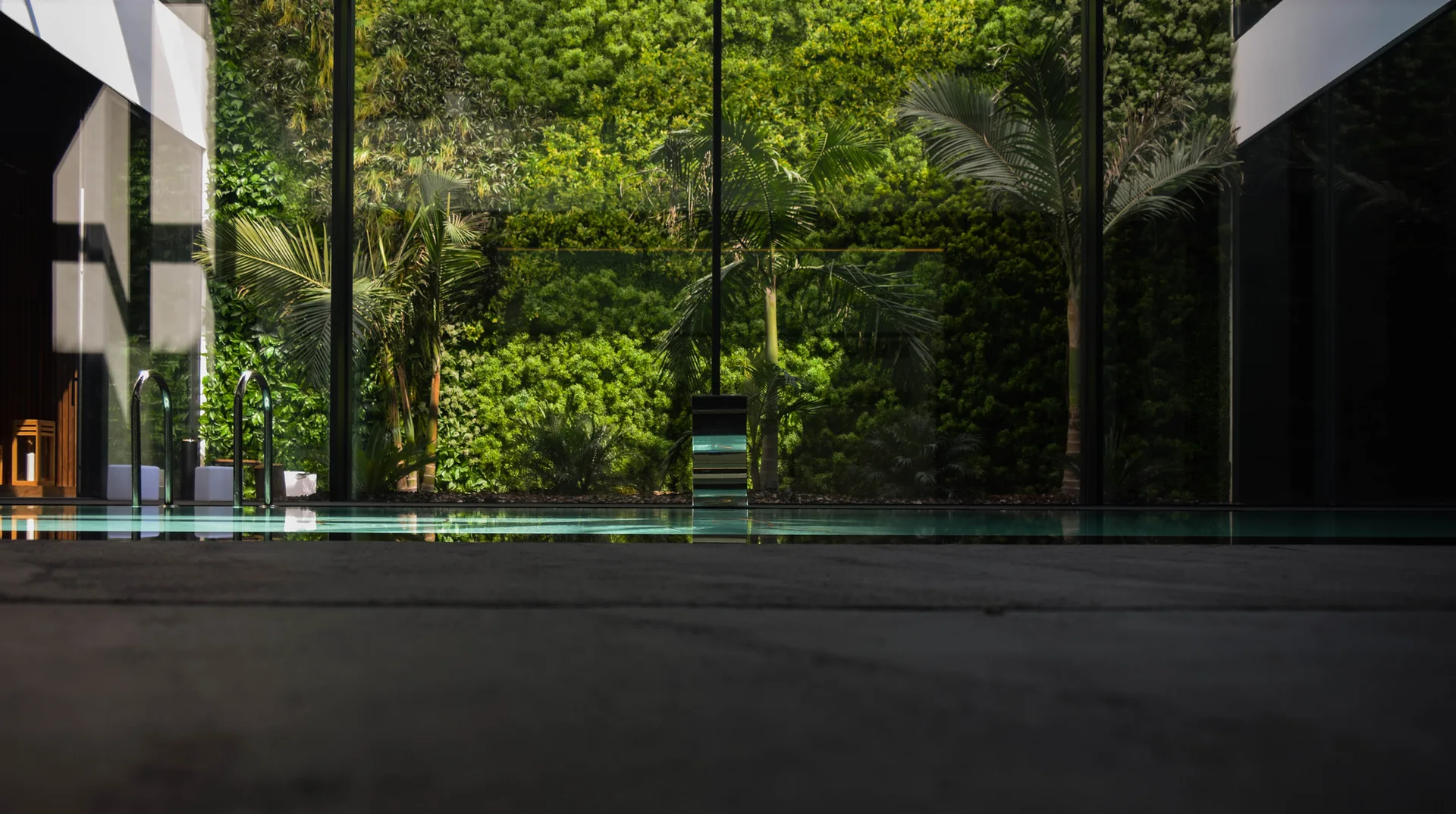 An indoor swimming pool features a metal ladder and a floor-to-ceiling glass window showcasing a towering outdoor vertical garden wall of lush green foliage.