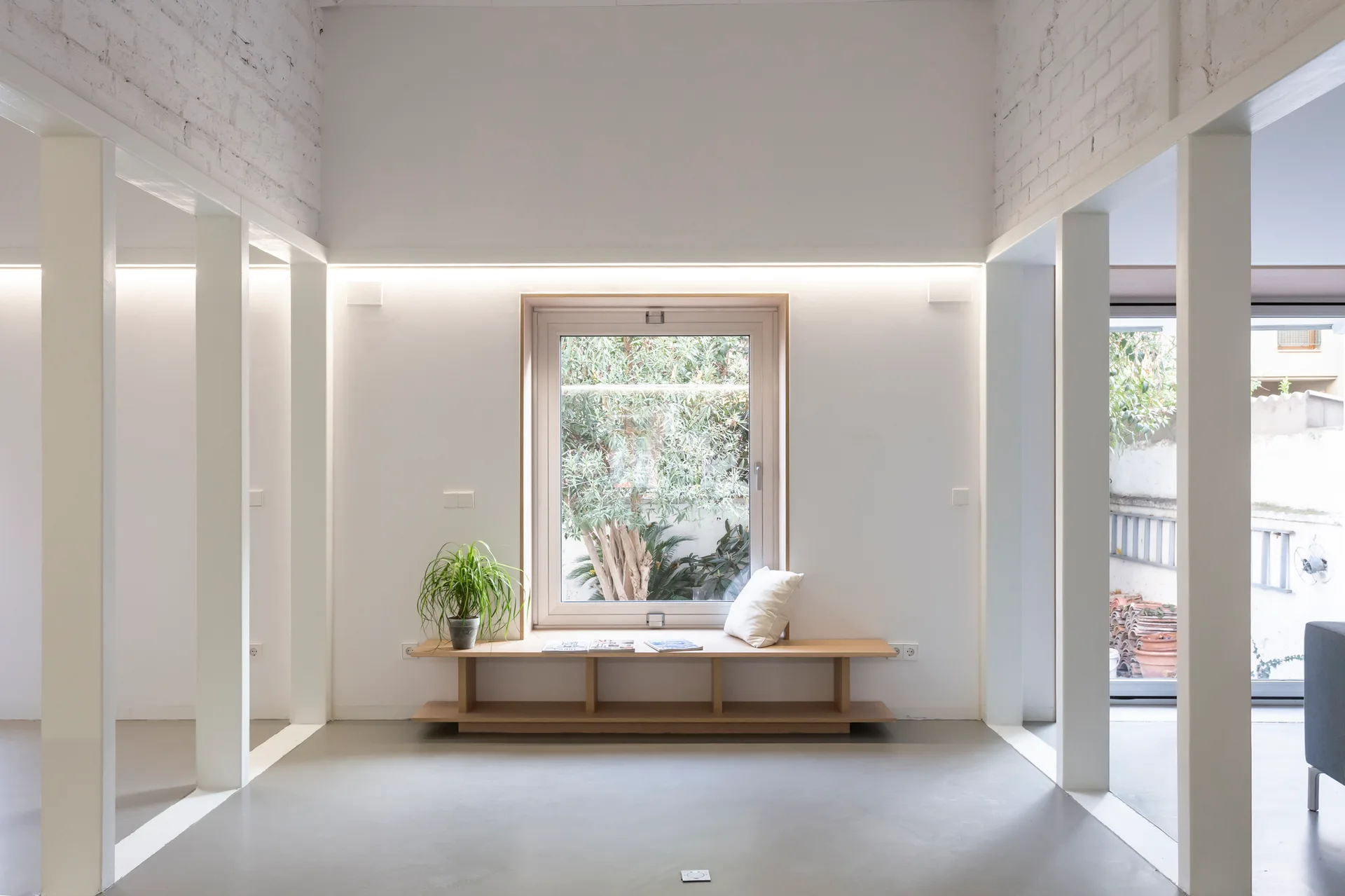 A minimalist white room features a low wooden window bench beneath a central window, flanked by white columns and lit by a linear cove light.