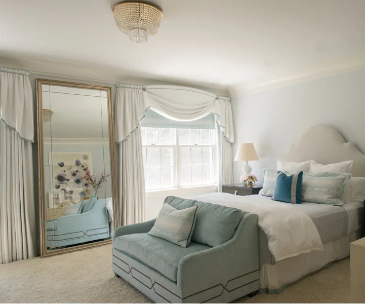 A light blue bedroom featuring a large paneled floor mirror next to a window with white drapes and an upholstered bed with a matching bench.