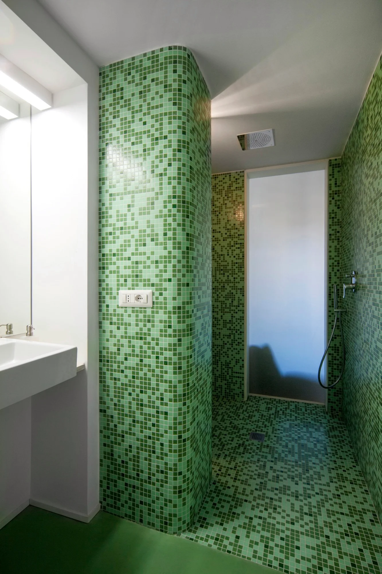 A modern bathroom featuring a curved wall and floor clad in green mosaic tiles next to a white sink and glass shower door.