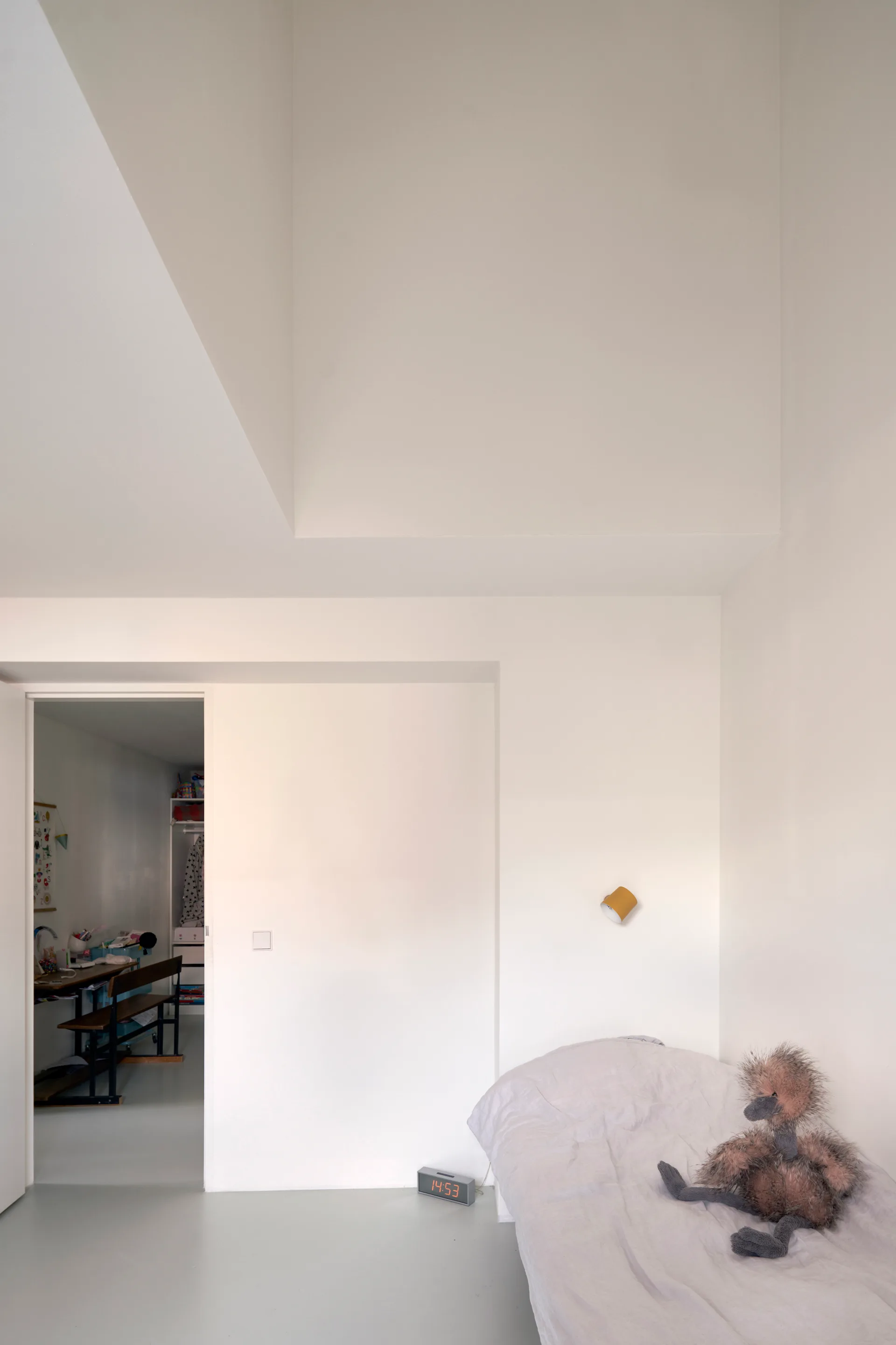 A minimalist white bedroom features a single bed with a plush toy, a gold wall sconce, and a doorway opening to an adjacent room.