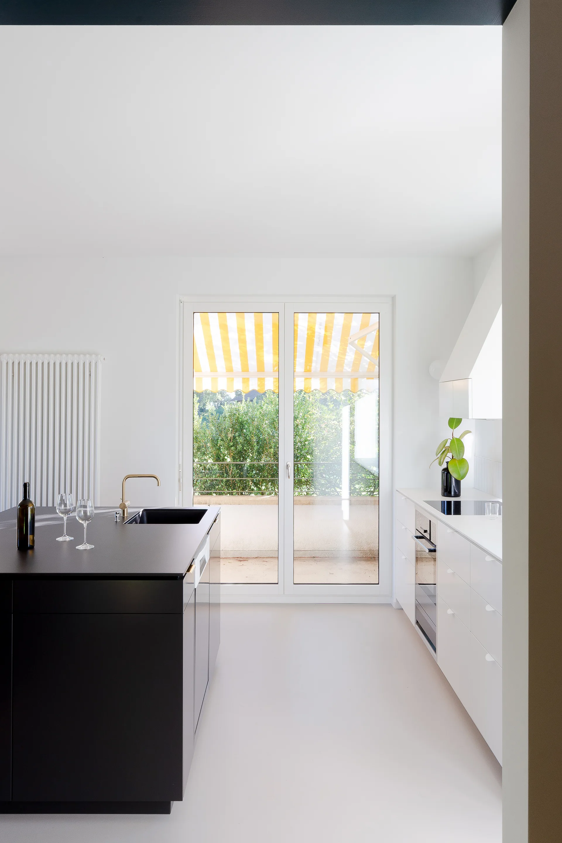 A minimalist kitchen featuring a matte black island with a gold faucet, white cabinets, and a glass door opening to a balcony with a yellow striped awning.