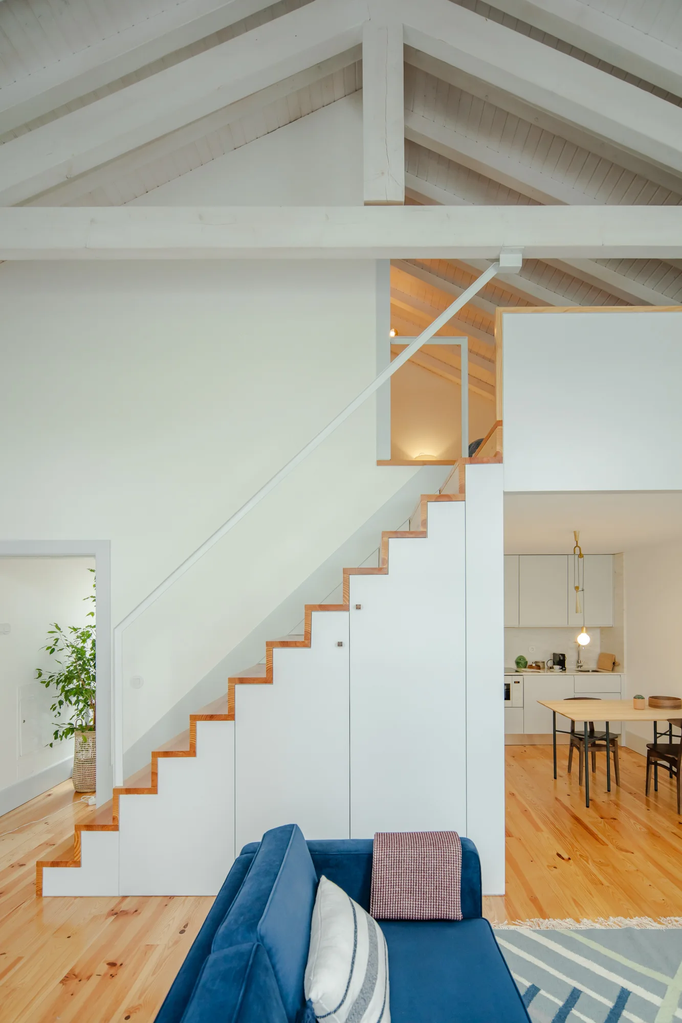 A modern open-plan living room with a white staircase featuring integrated storage, a blue sofa, and exposed ceiling beams.