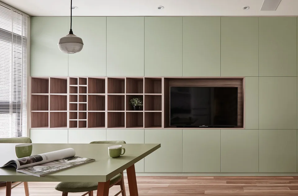A minimalist dining room features a sage green storage wall with a wood-lined shelving niche and a matching green dining table.