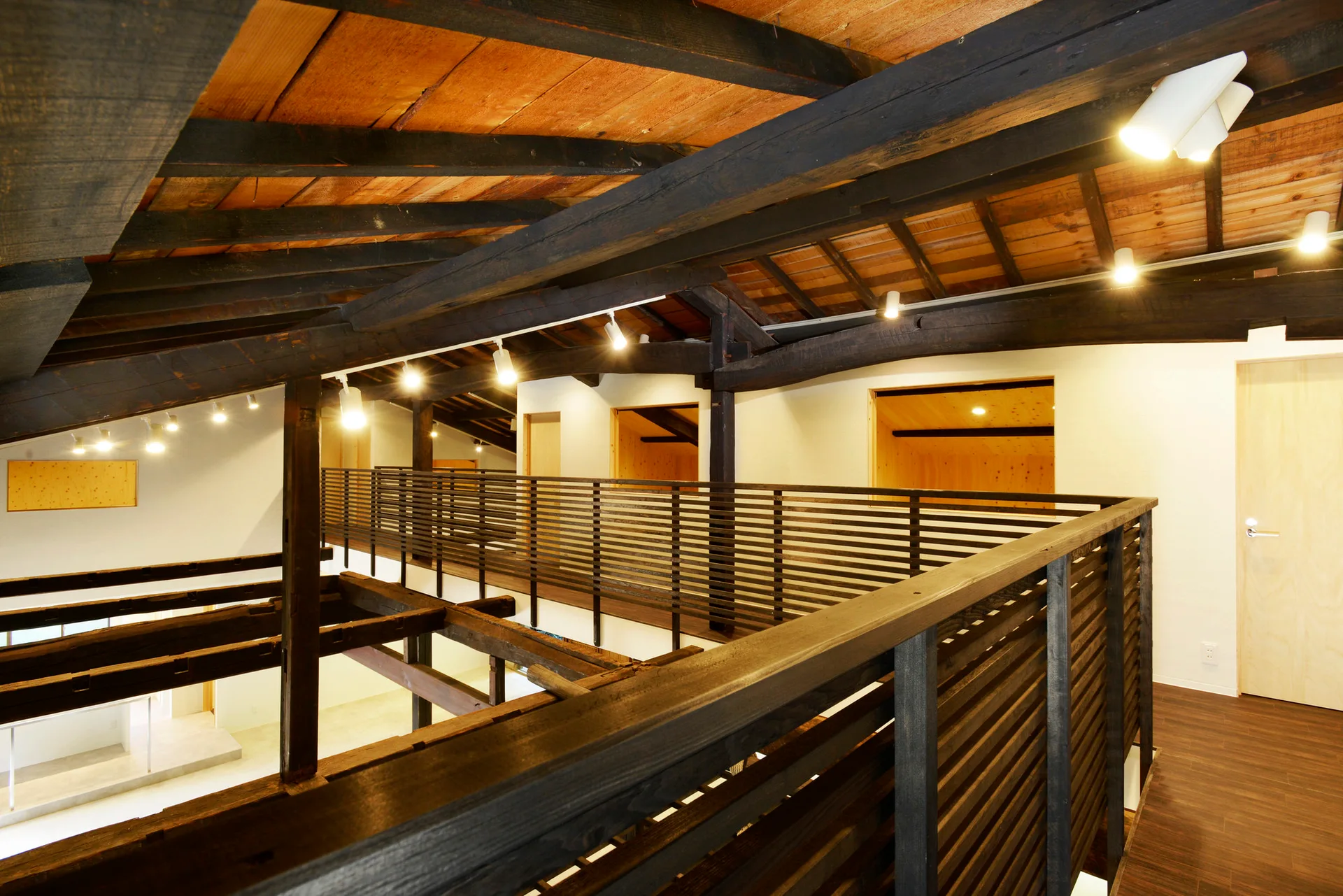 An elevated wood mezzanine walkway features a slatted railing beneath a pitched ceiling with dark exposed timber beams and spotlight track lighting.